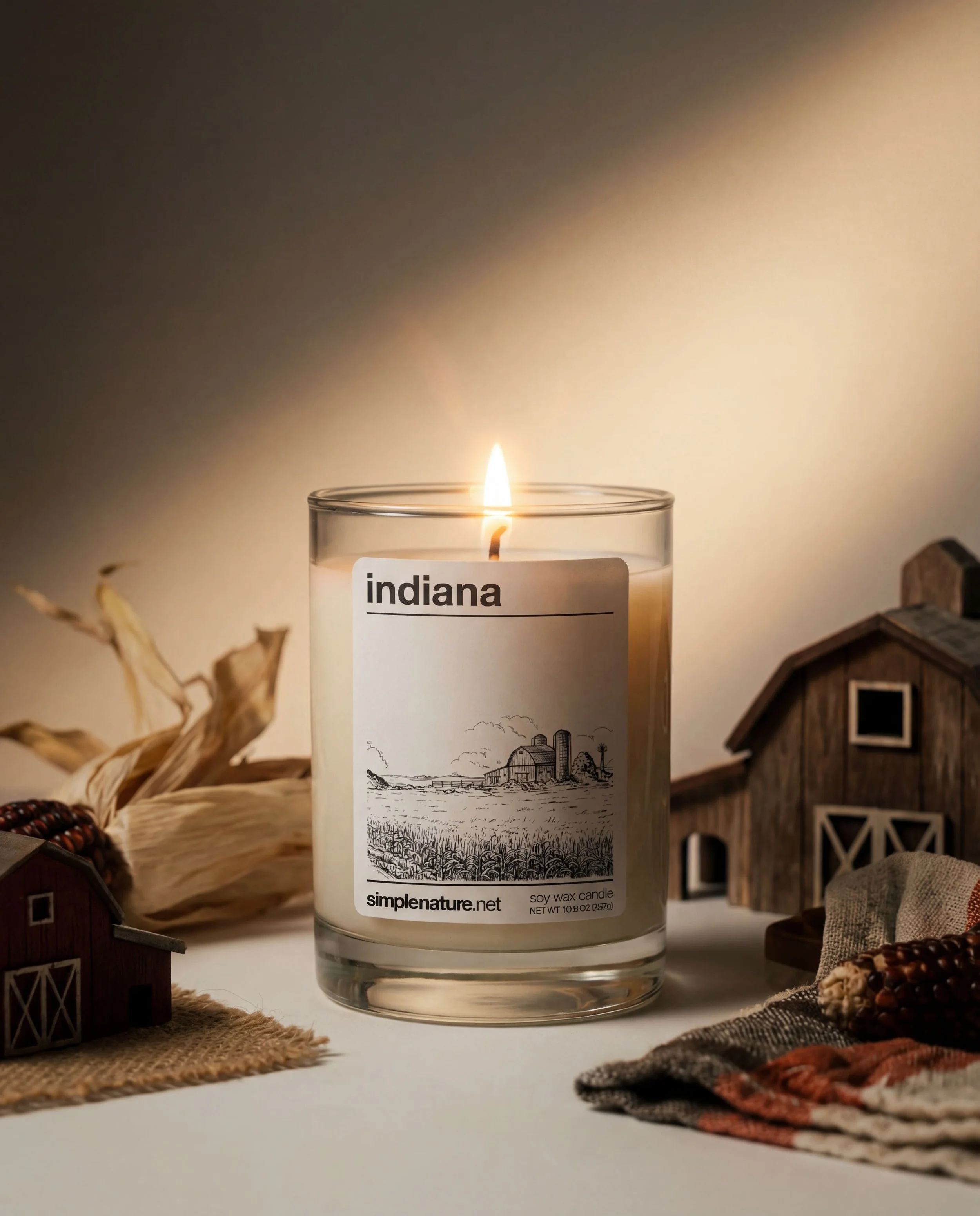 A lit soy wax candle with Indiana on the label, surrounded by small wooden farm structures and fabric.