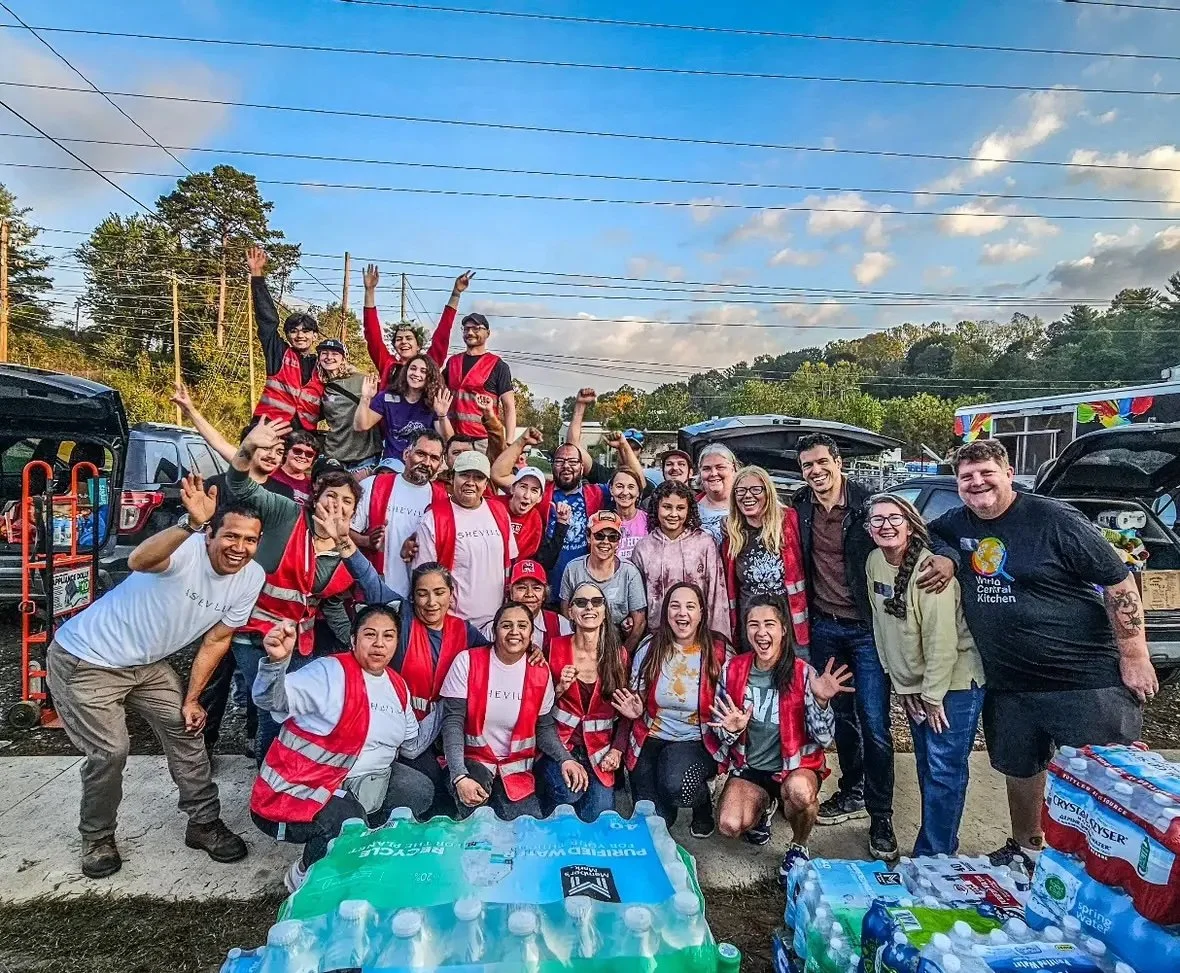 Group of diverse people celebrating outdoors, some wearing vests, surrounded by water bottles, with trees and cars in the background.