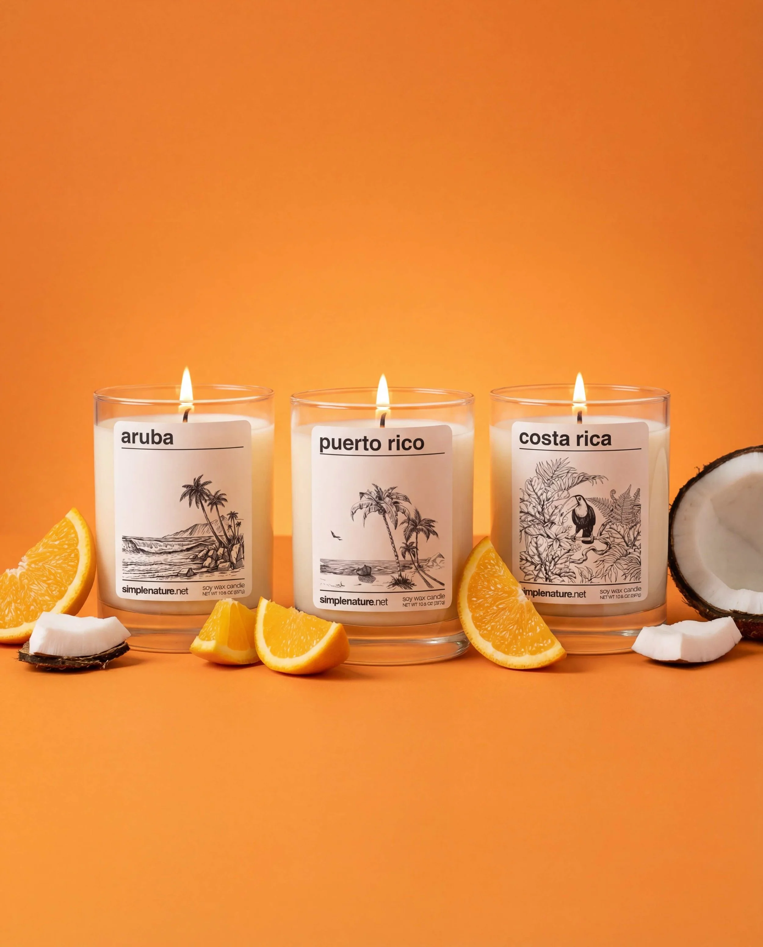 Three lit candles with tropical island labels, surrounded by sliced oranges, lemons, and pineapple on sand.