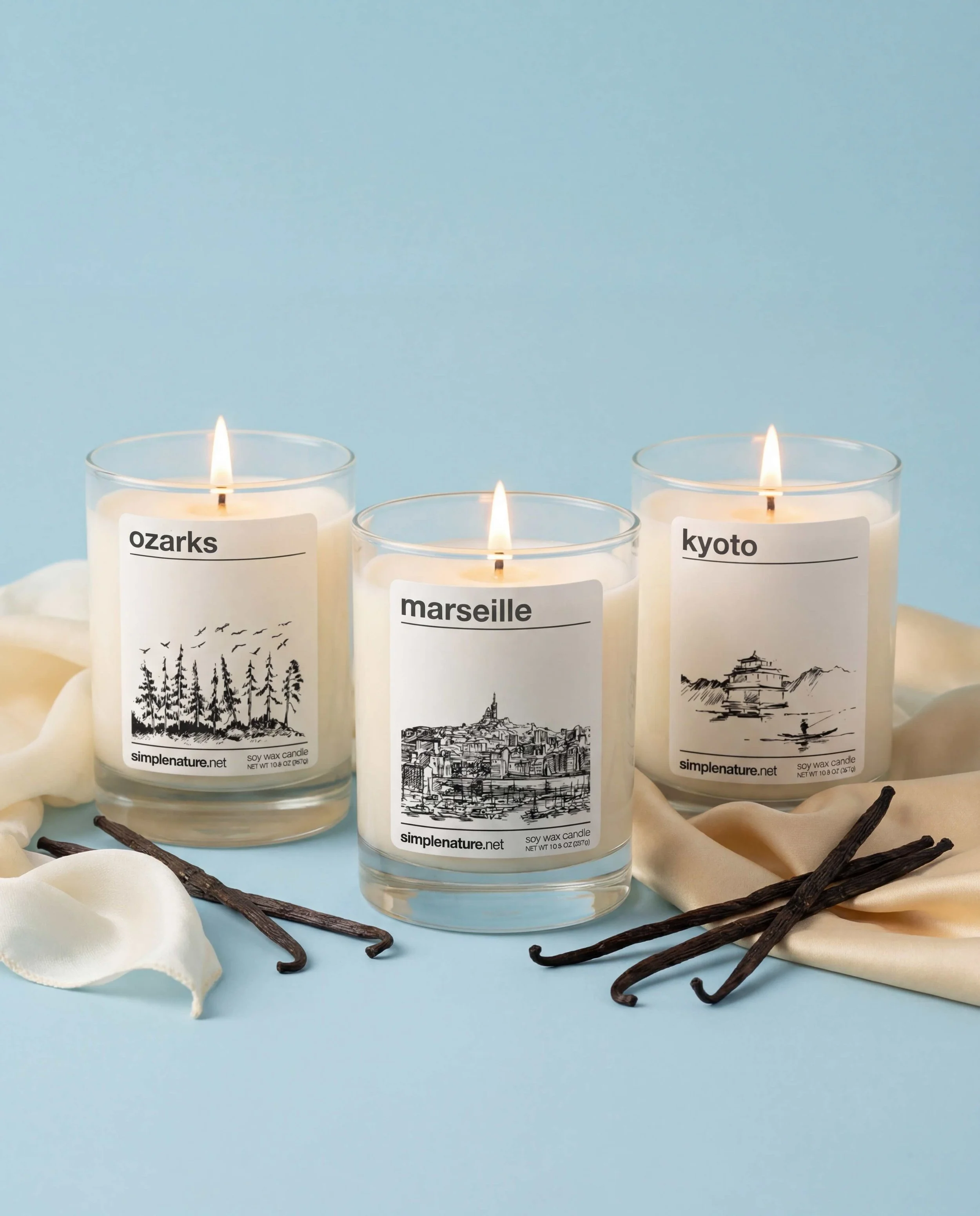 Three lit candles in glass containers with nature-themed labels reading 'aurora,' 'explore,' and 'golden moon'; they are on a textured white surface with a metallic blue corrugated background.