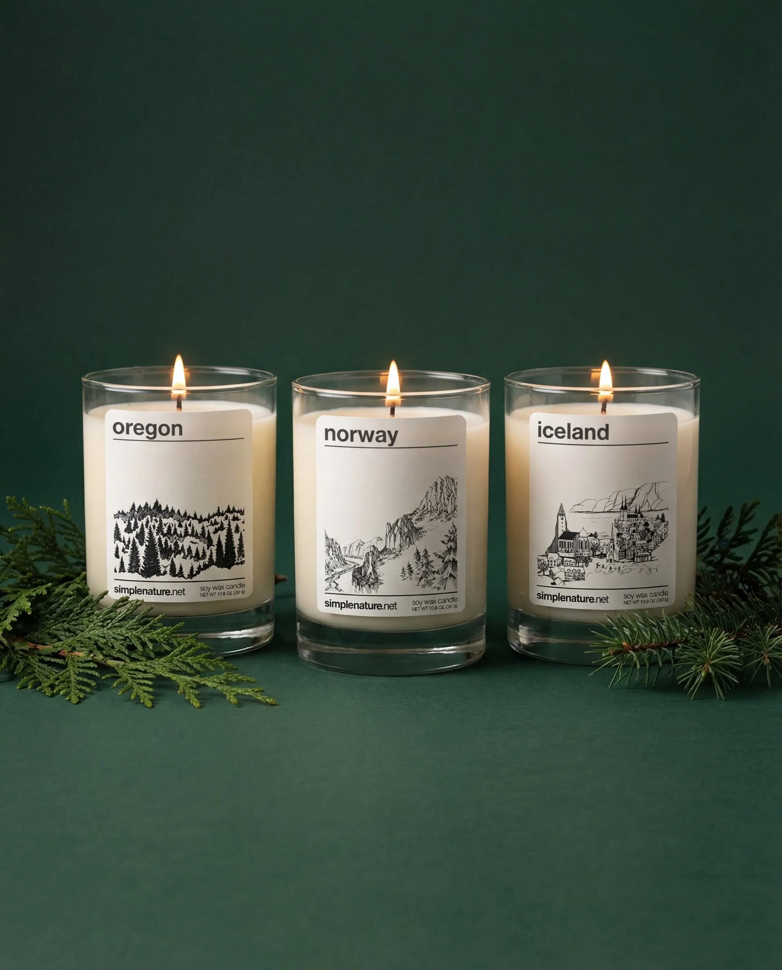 Two lit candles with labels 'Norway' and 'Oregon' on a wooden surface surrounded by pine branches and logs, with a dark background.