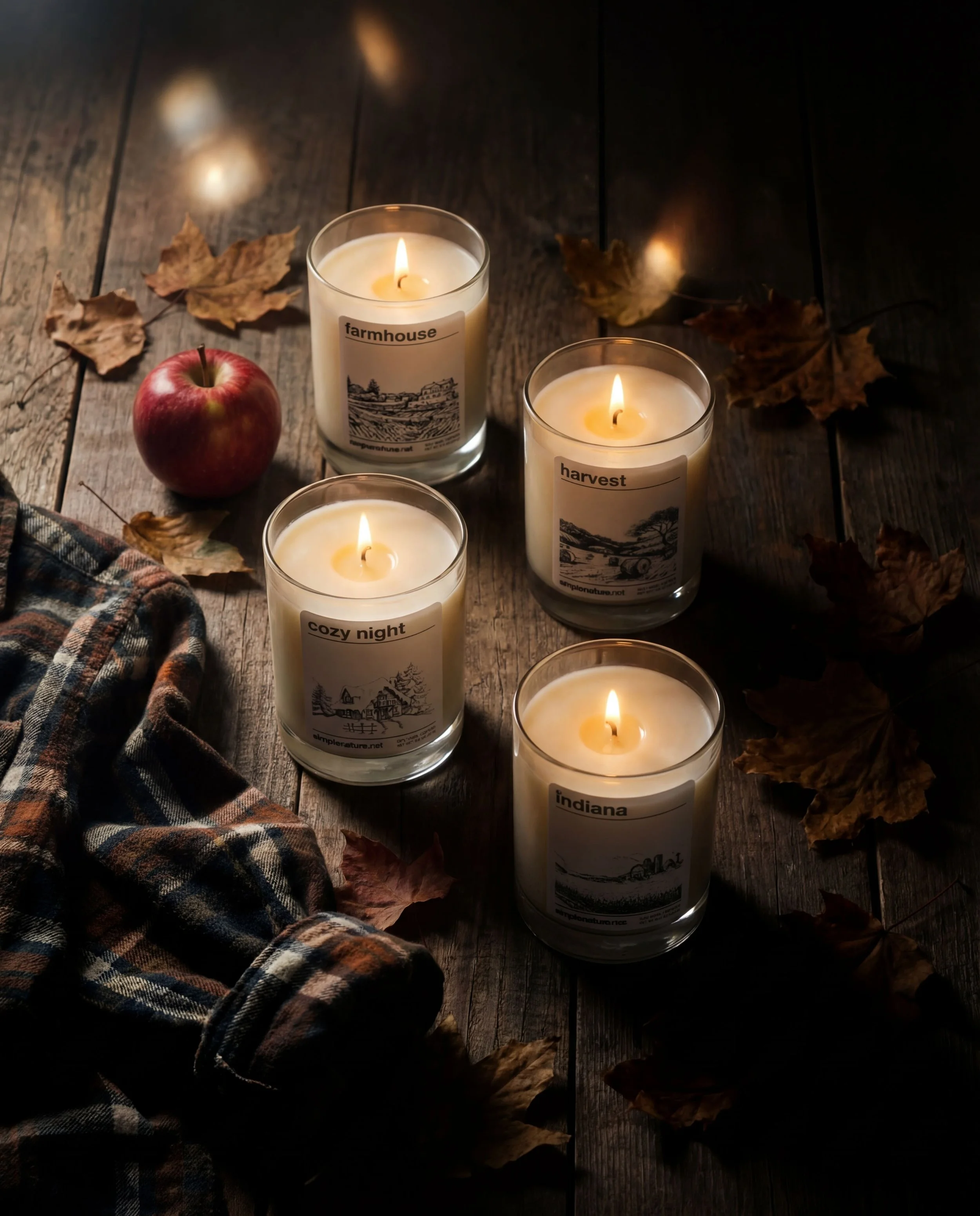 Four lit candles in glass holders with seasonal labels, a red apple, a plaid cloth, and scattered dry autumn leaves on a wooden surface.