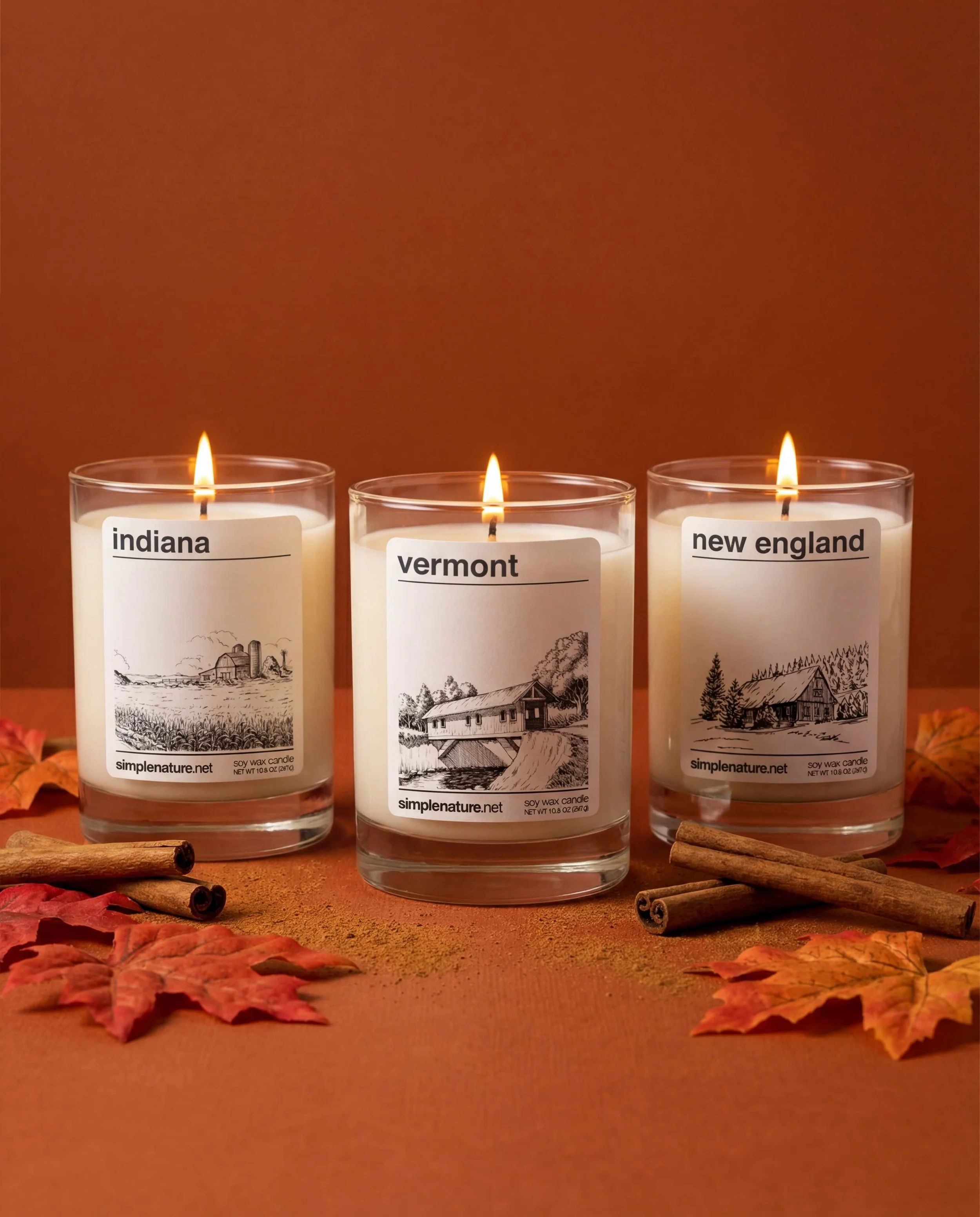 Four lit candles in glass holders with seasonal labels, a red apple, a plaid cloth, and scattered dry autumn leaves on a wooden surface.