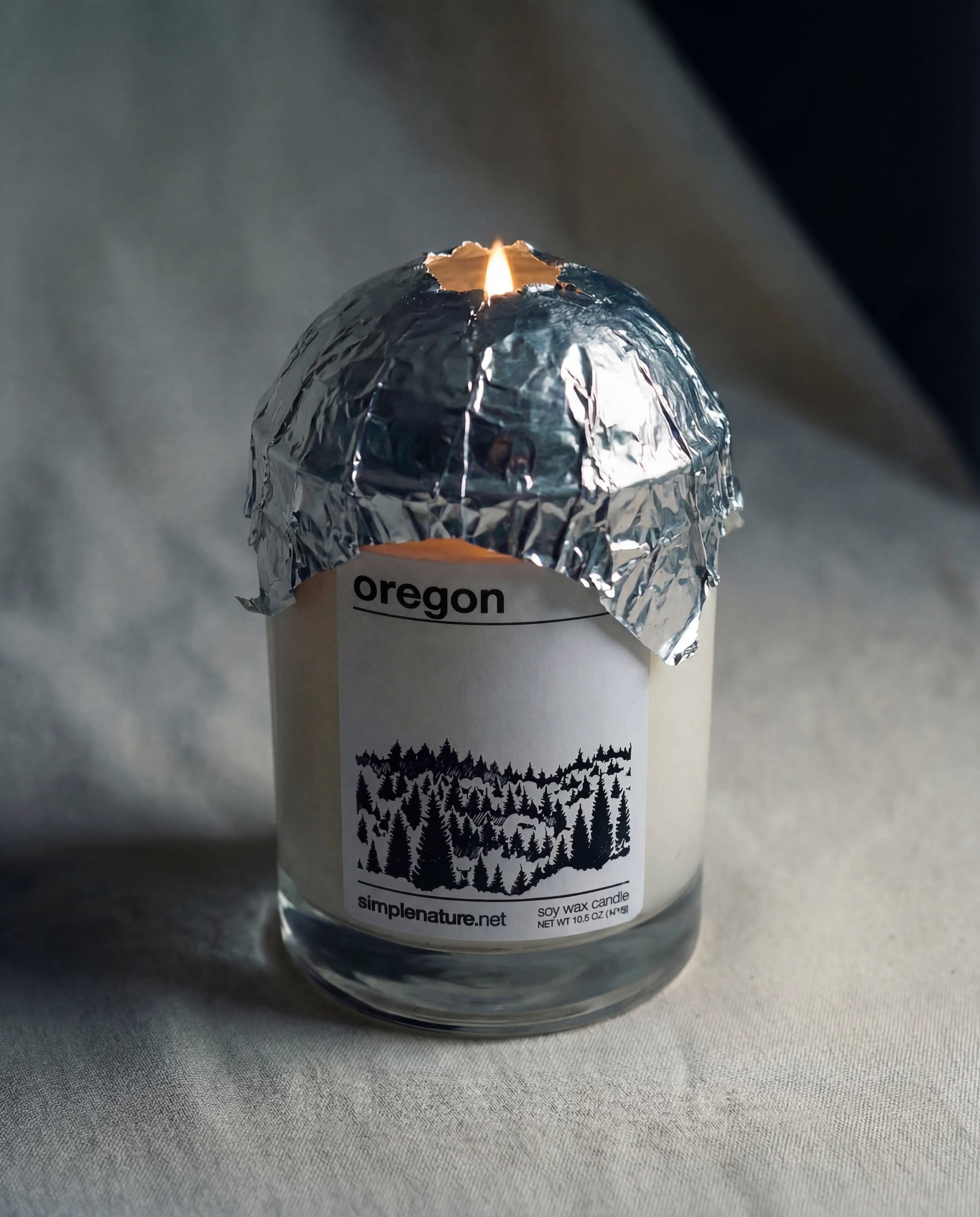 A soy wax candle in a glass jar with a tin foil lid partially removed, lit and burning at the top. The candle label indicates it is from Oregon and features a forest illustration.