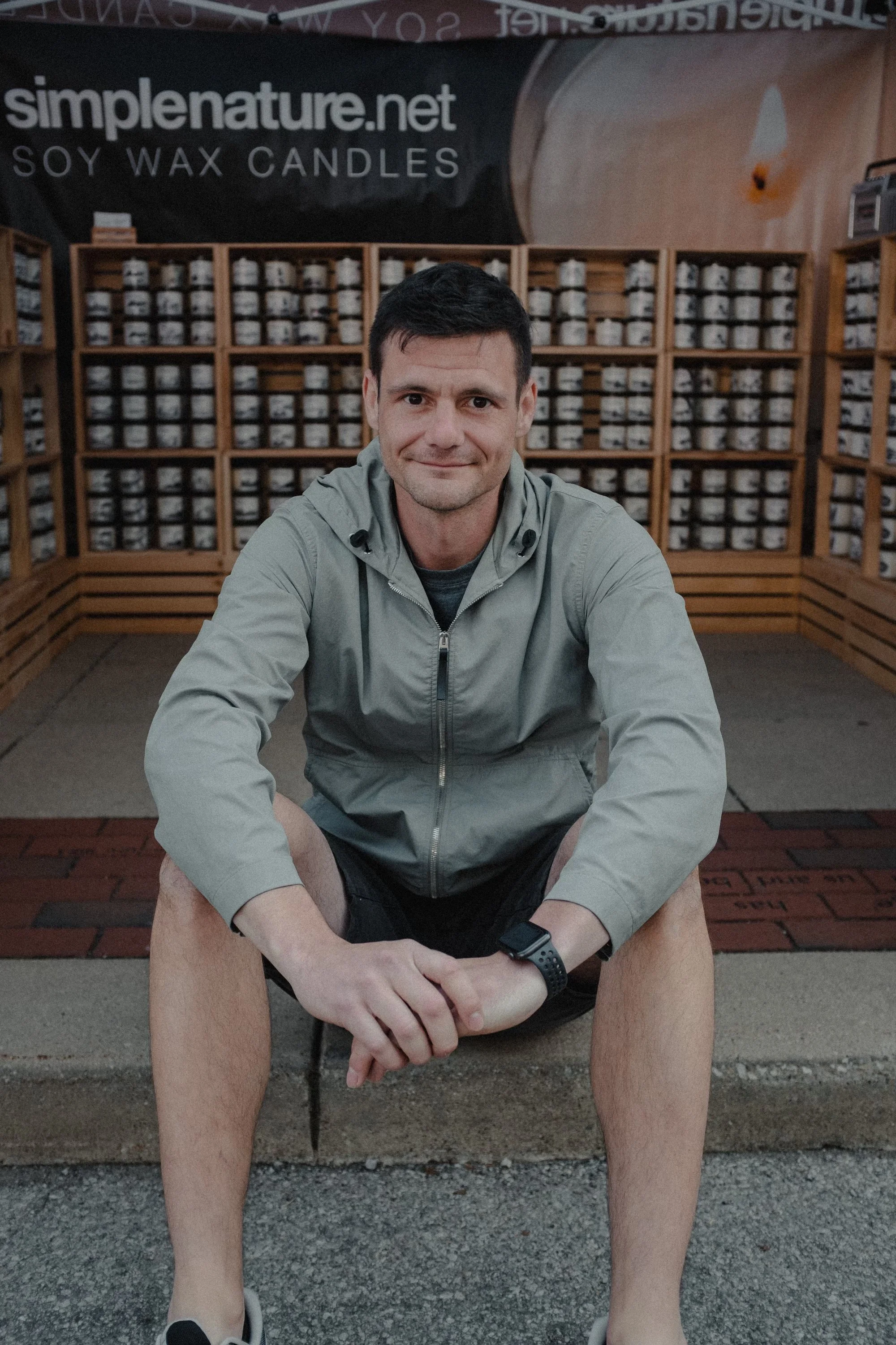 A man sitting on a curb in front of shelves filled with candles and a sign for 'simplenature.net soy wax candles'