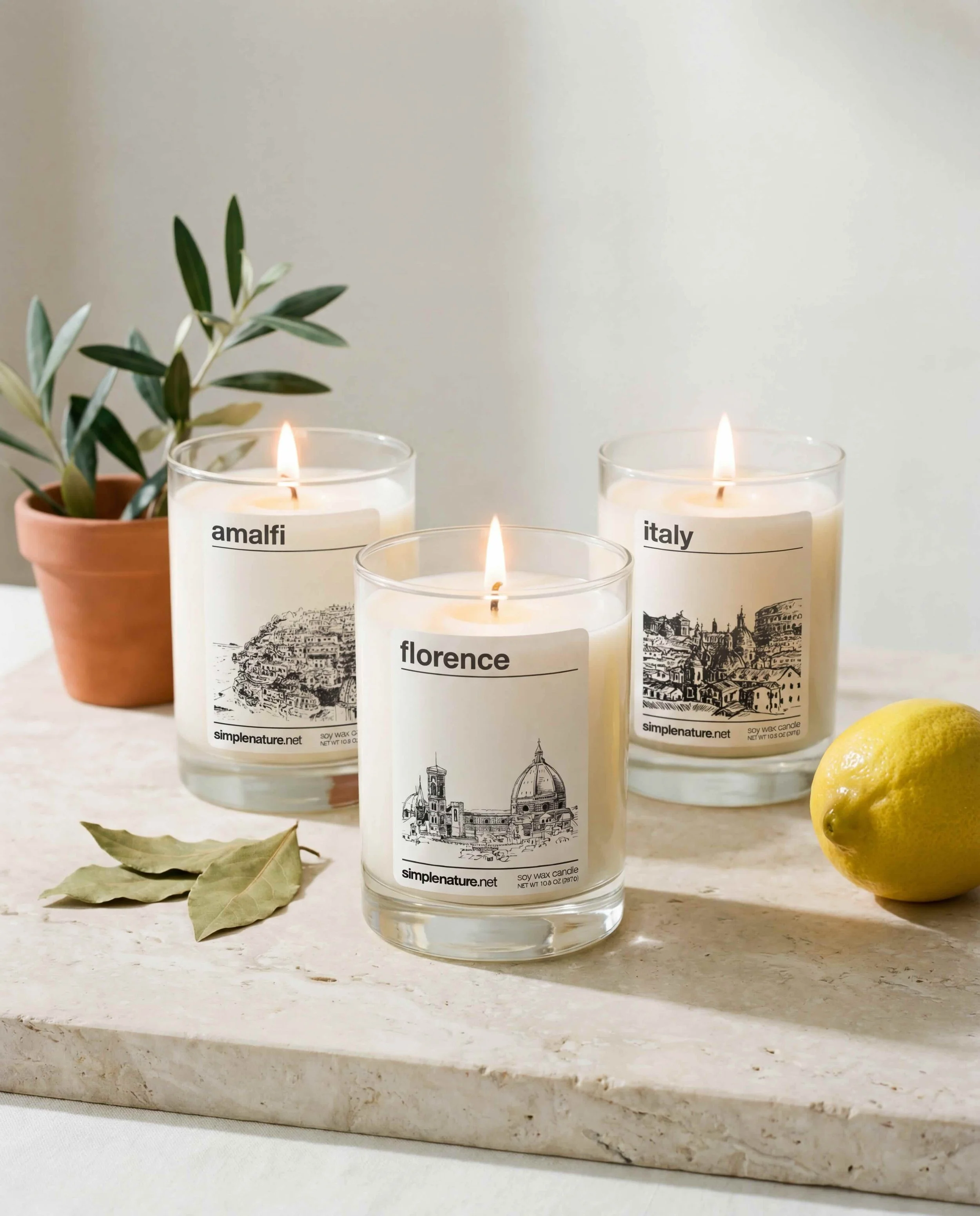 Three lit candles with labels of Florence, Italy, and Amalfi, placed on a marble surface with a potted plant, a lemon, and dried leaves nearby.