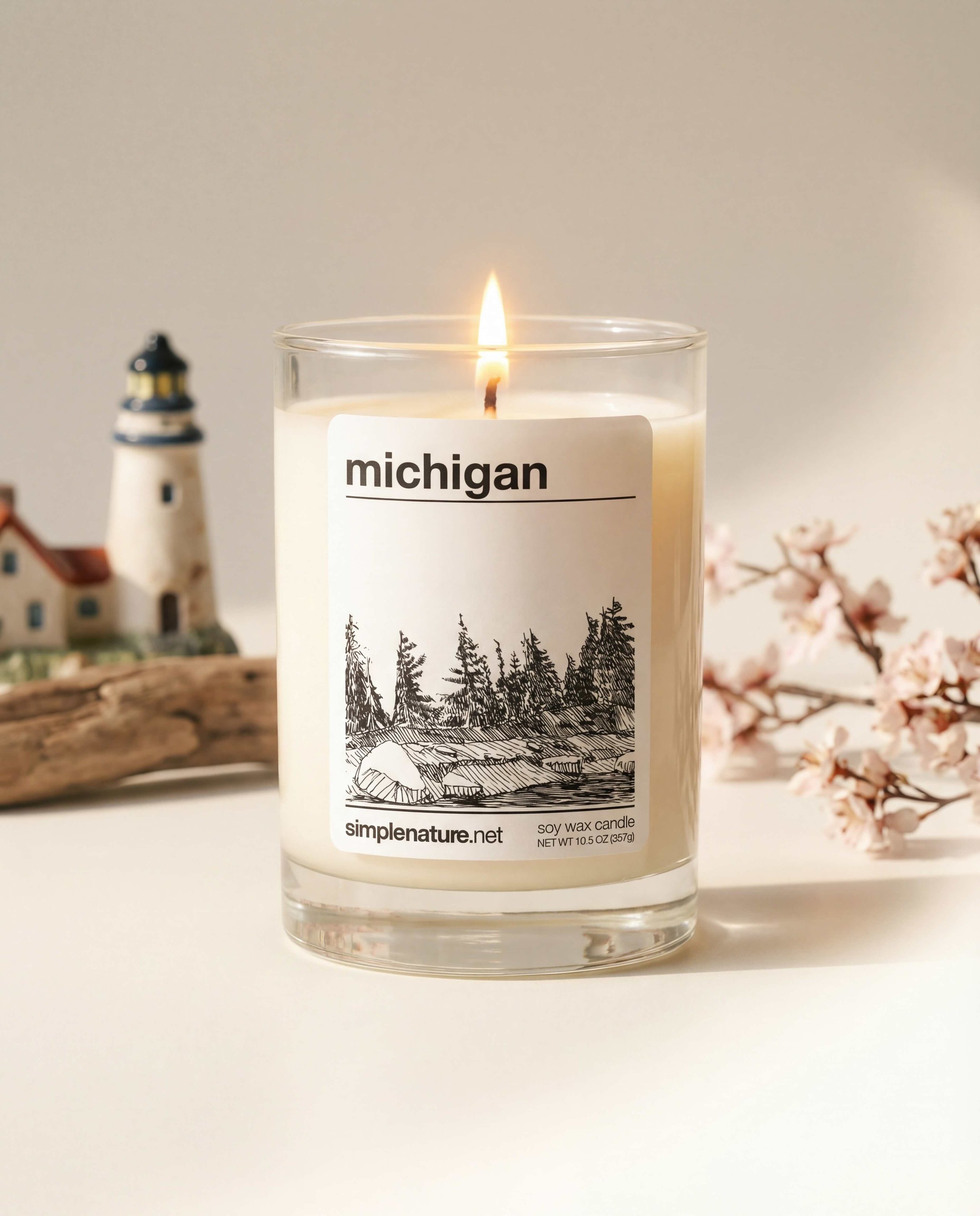 A lit soy wax candle in a clear glass container with a label that reads "michigan" and features a black and white illustration of trees and rocks. In the background, there is a model lighthouse, a miniature house, a piece of driftwood, and pink cherry blossom branches.