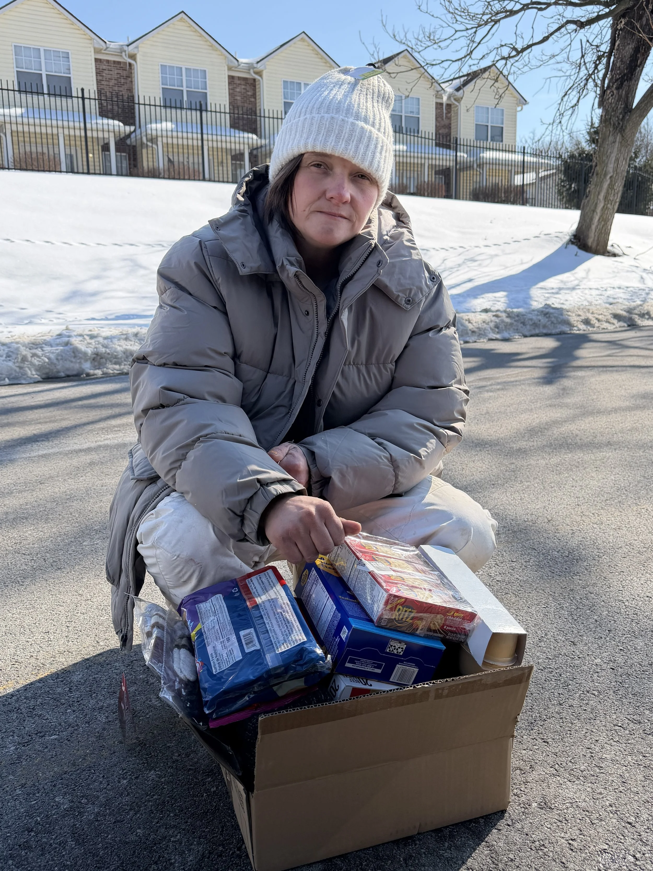 A woman wearing a white knit hat and gray puffy jacket kneeling on a paved street next to a cardboard box filled with various grocery items, with snow on the ground and a row of yellow and brick apartment buildings in the background.