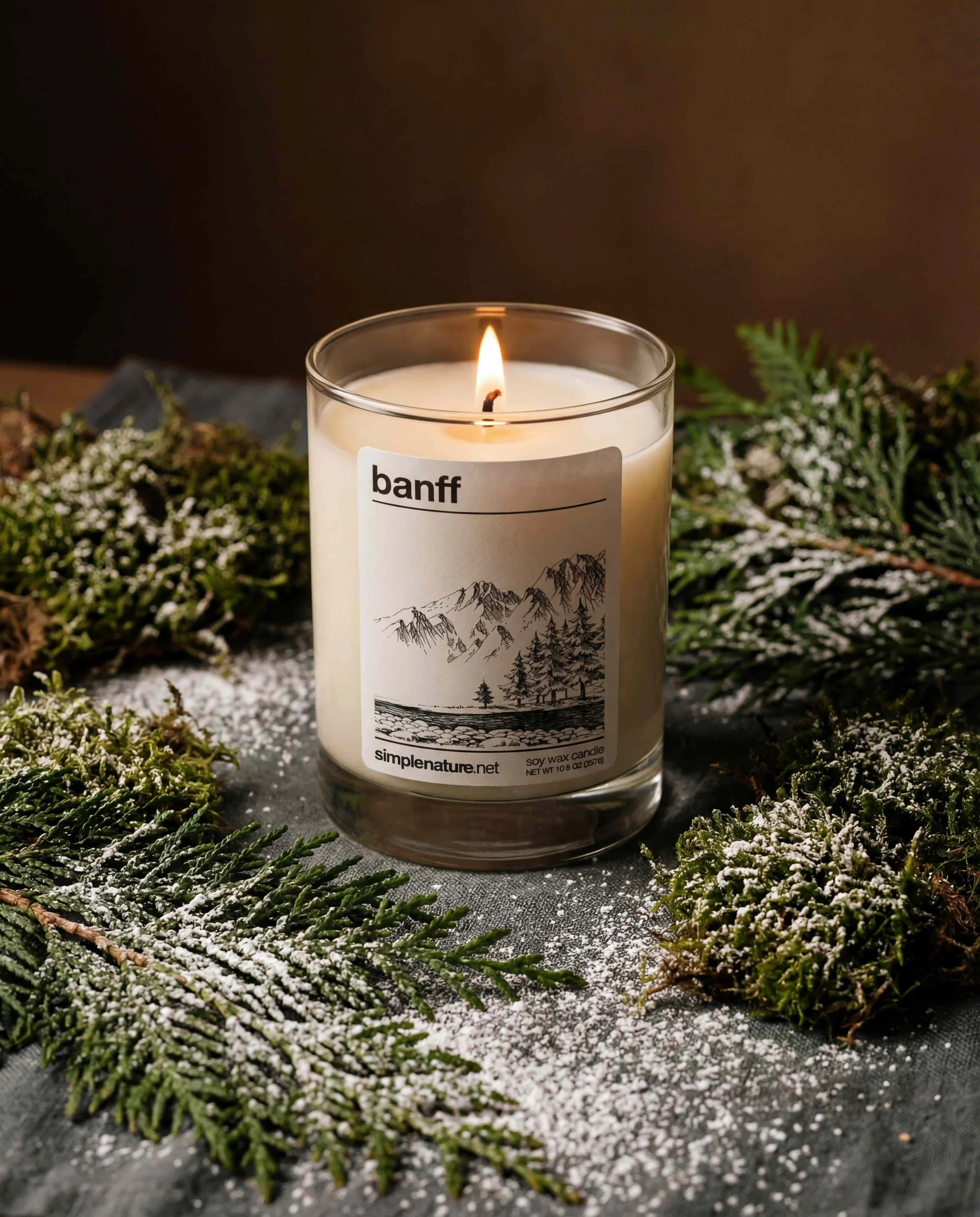 A lit candle in a glass jar labeled 'Banff' surrounded by pine branches dusted with snow.