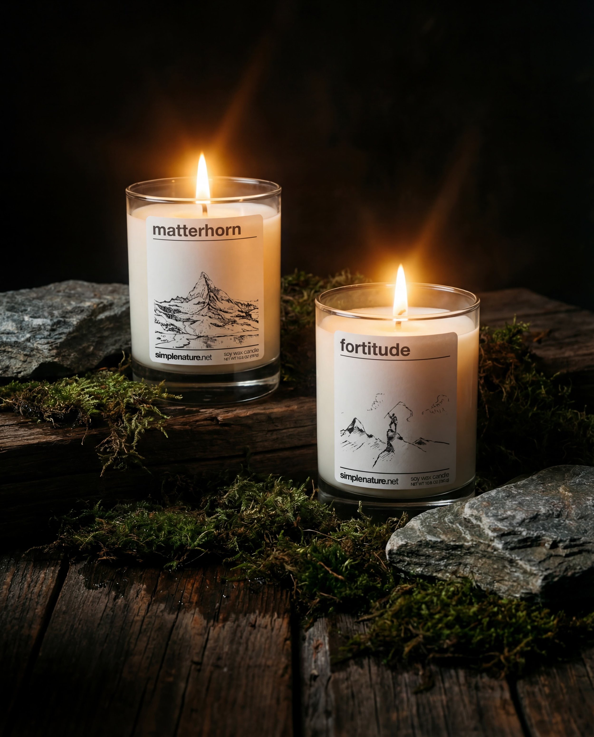 Two lit candles in glass holders placed on a wooden surface with moss and rocks. Each candle has a label with the words 'matterhorn' and 'fortitude,' along with line drawings of mountain scenes.