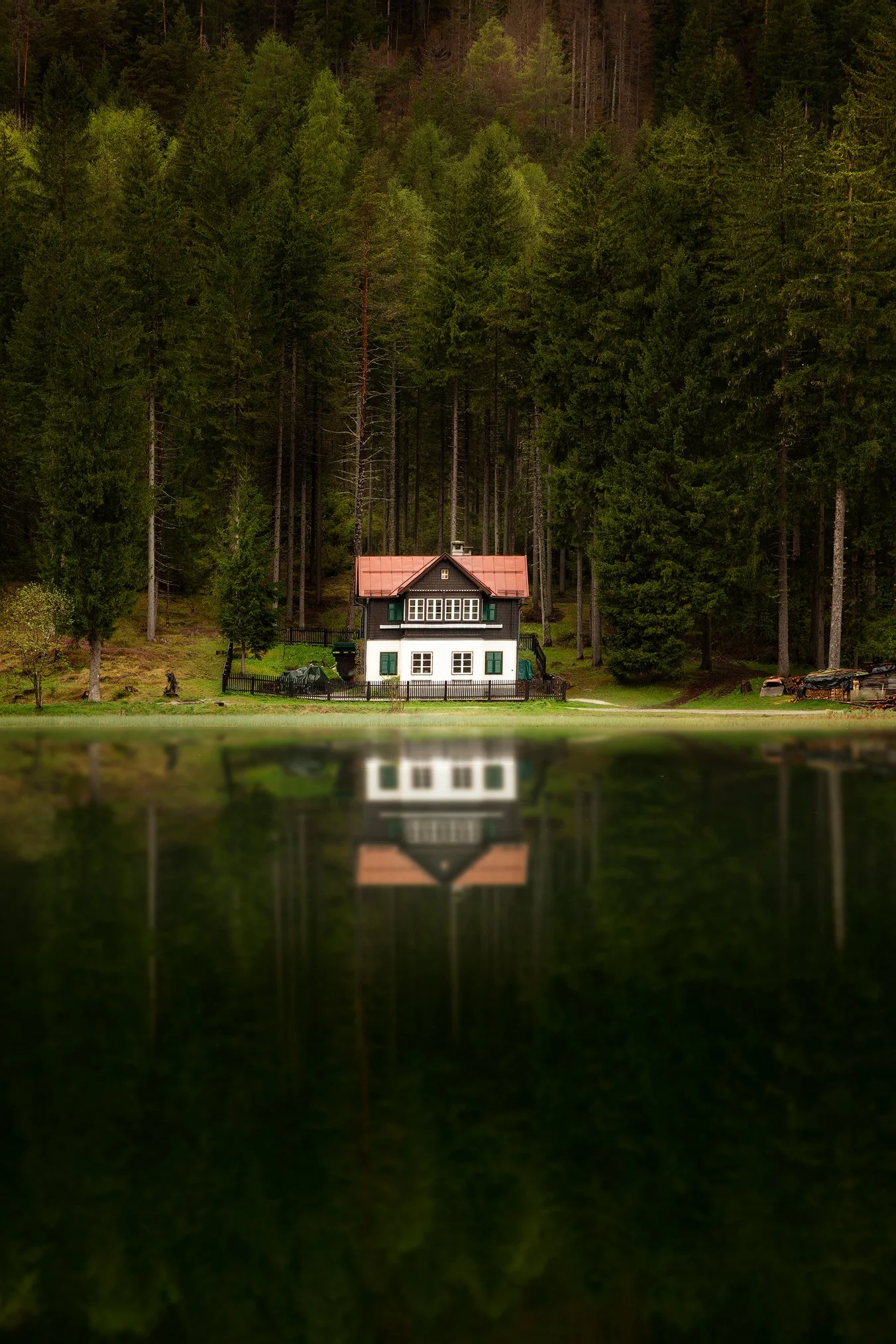 Une maison située au bord d'un lac, entourée de grands arbres verts, avec leur reflet visible dans l'eau calme du lac.