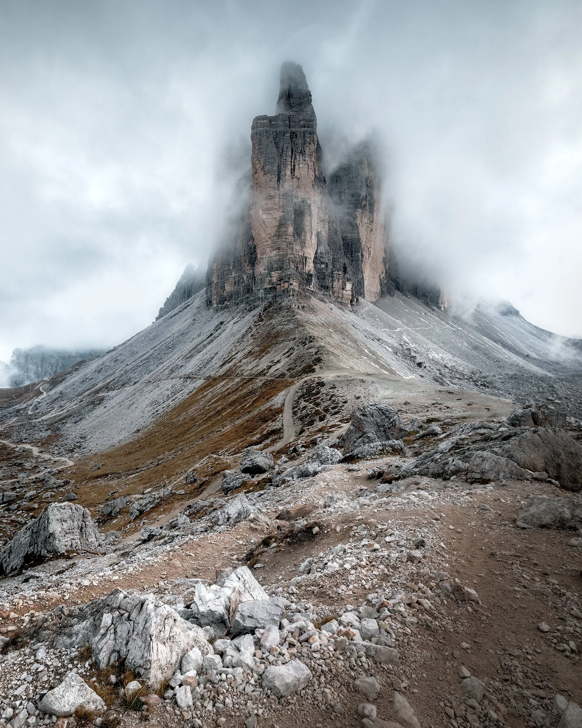 Une montagne rocheuse imposante entourée de nuages et de brouillard, avec un chemin de terre sinueux au premier plan.