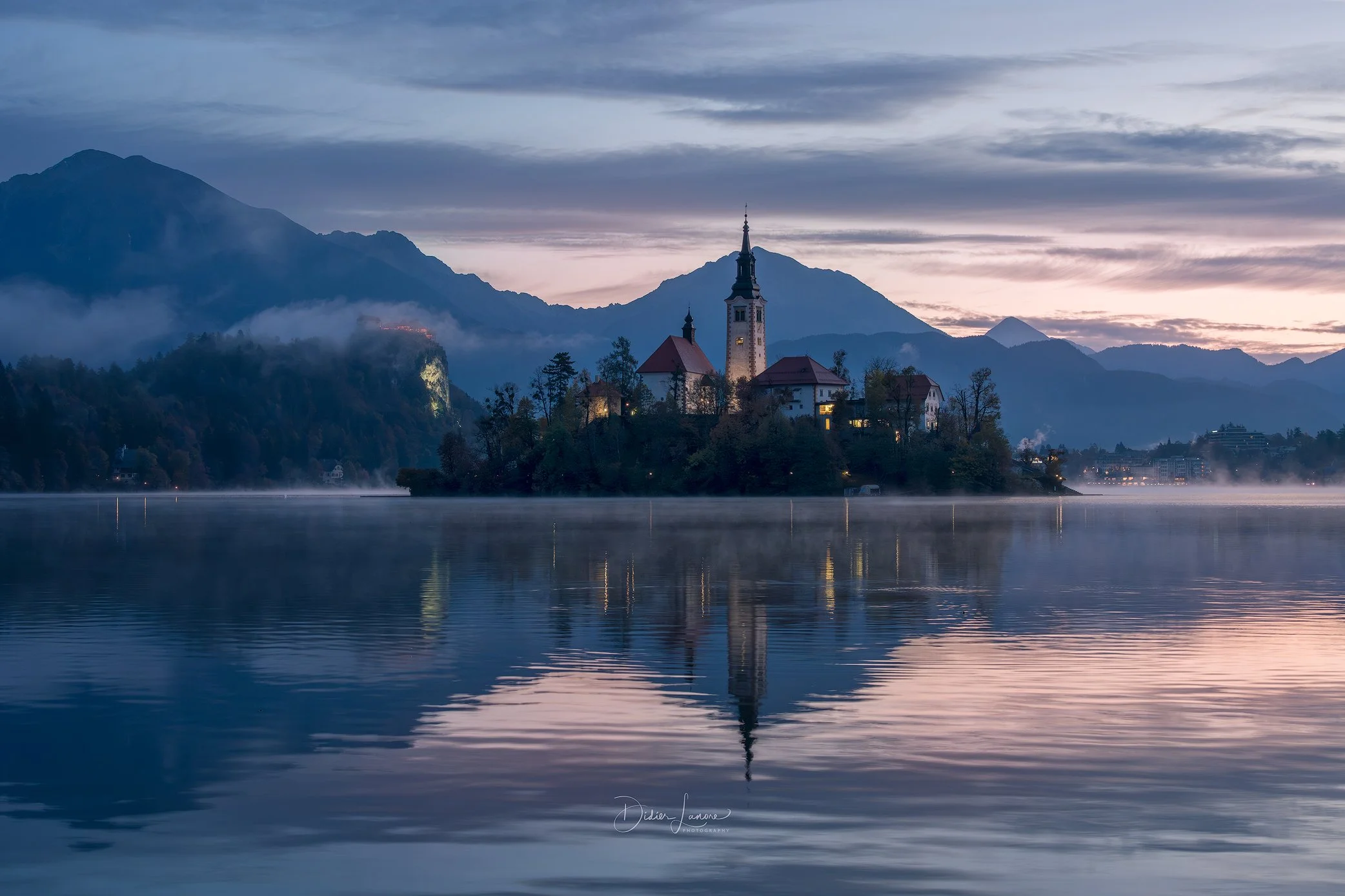 Une île avec une église et des bâtiments entourés d'eau calme, avec des montagnes en arrière-plan au lever ou coucher du soleil.