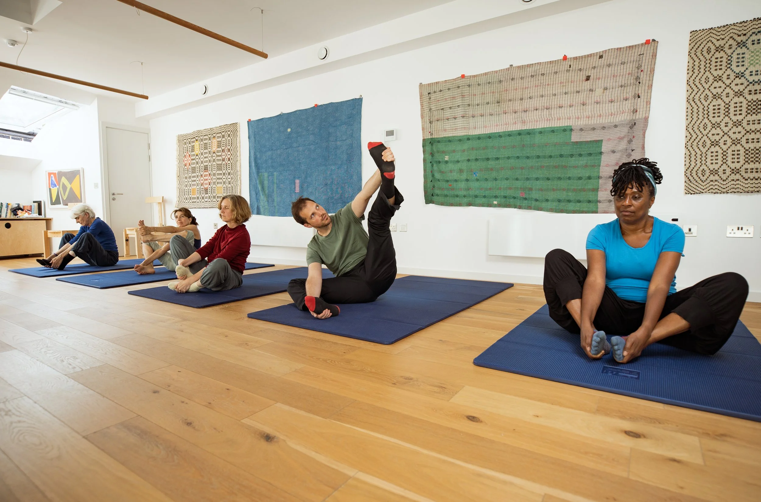 A group of people of various ages performing seated Feldenkrais exercises on blue mats in a bright room with wooden floors and colourful wall hangings.