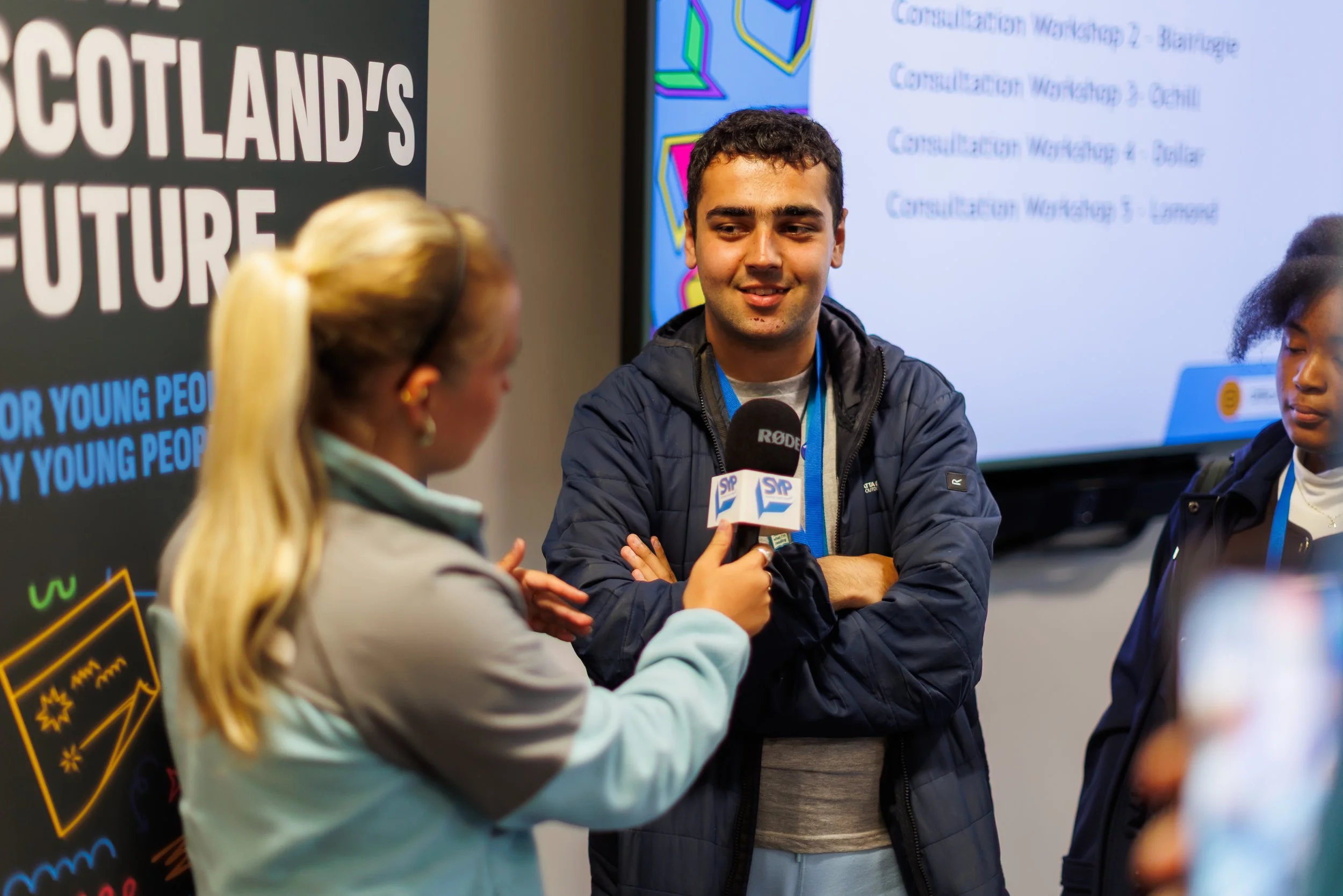 An MSYP with blond heair and blue jacket holding a mic and interviewing another MSYP wearing a dark blur jacket