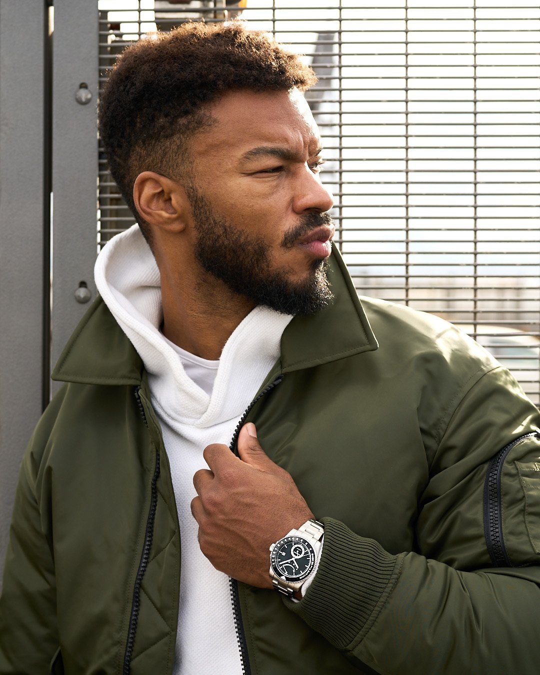 In a green bomber jacket and with a black dial pulsometer men’s stylish watch on his wrist, a black male model leans against a fence and gazes sideways