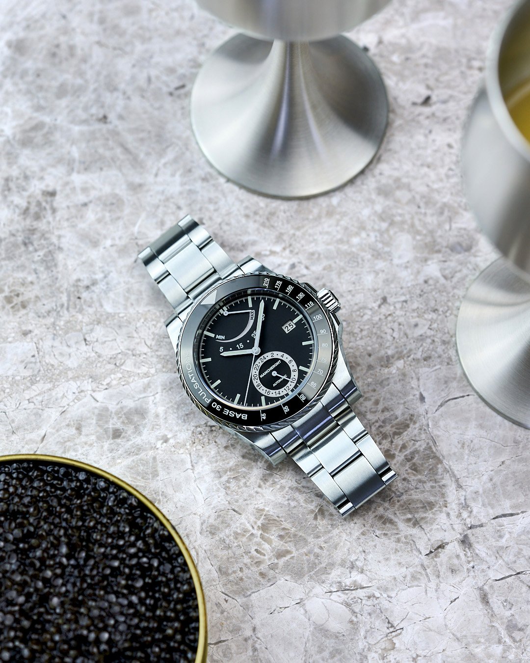 Black dial pulsometer men’s formal watch with stainless steel bracelet on marble surface in between caviar, and pewter goblets with wine
