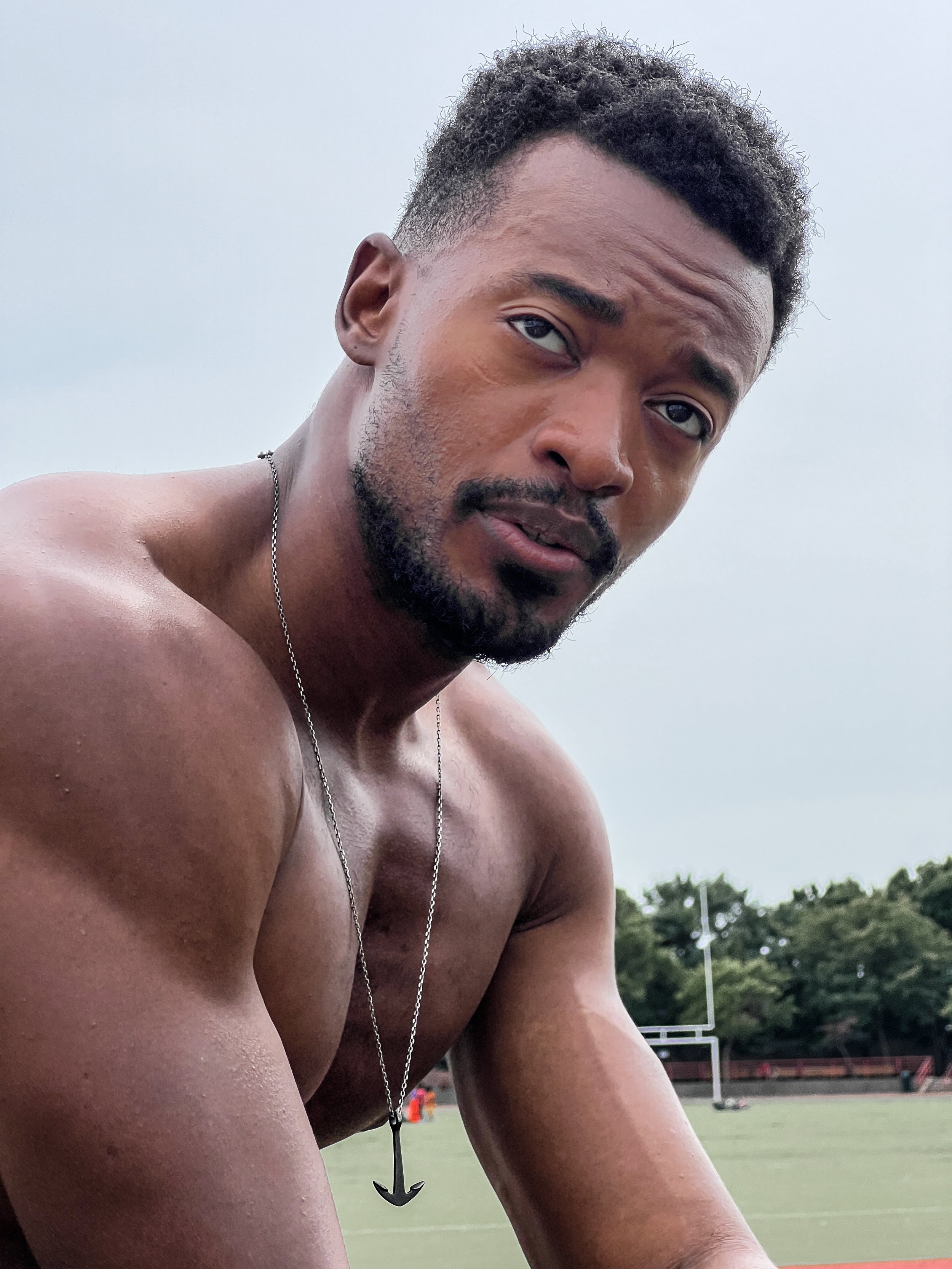Shirtless man with anchor necklace outdoors, athletic build.