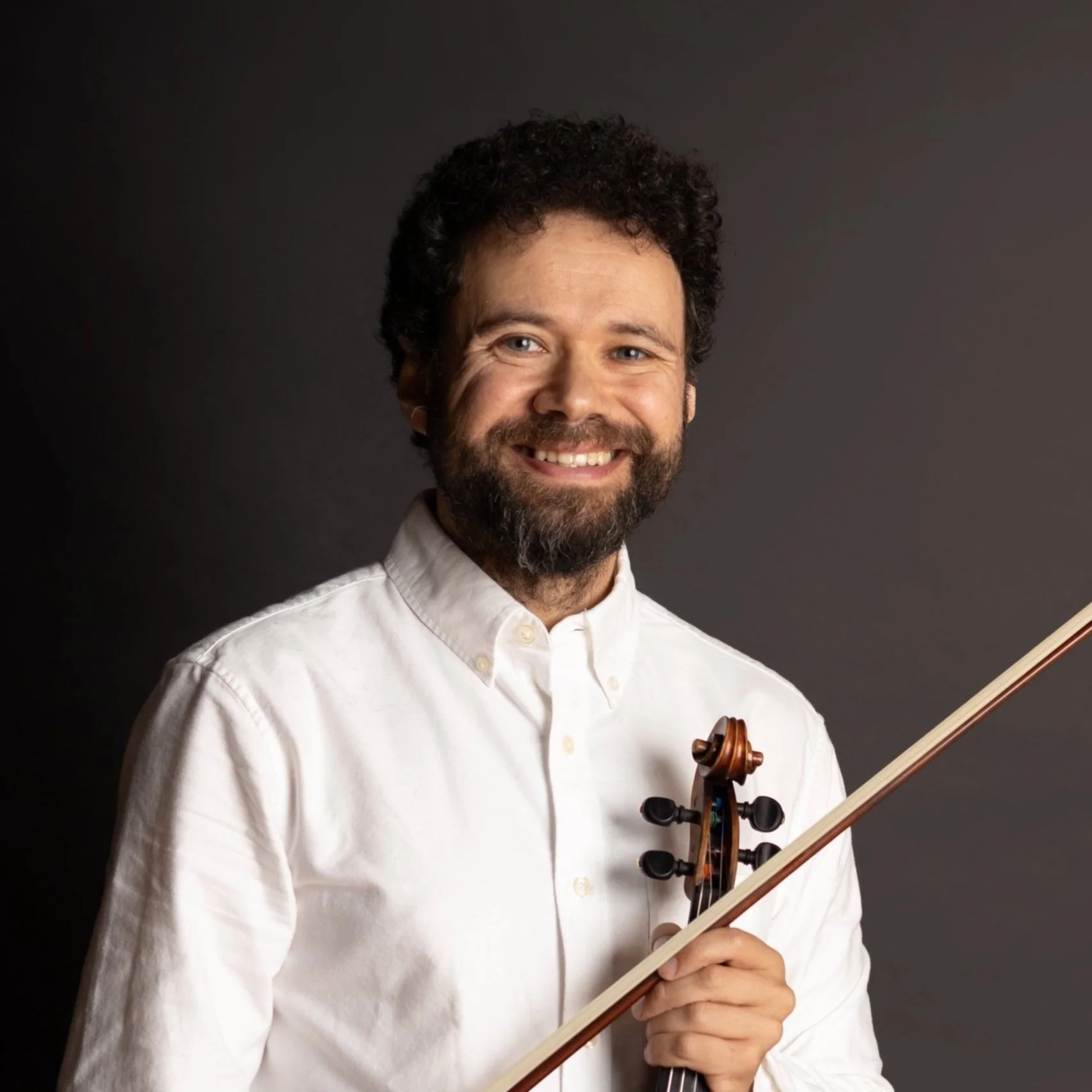 A man with curly dark hair and a beard smiling while holding a violin and bow against a dark background.