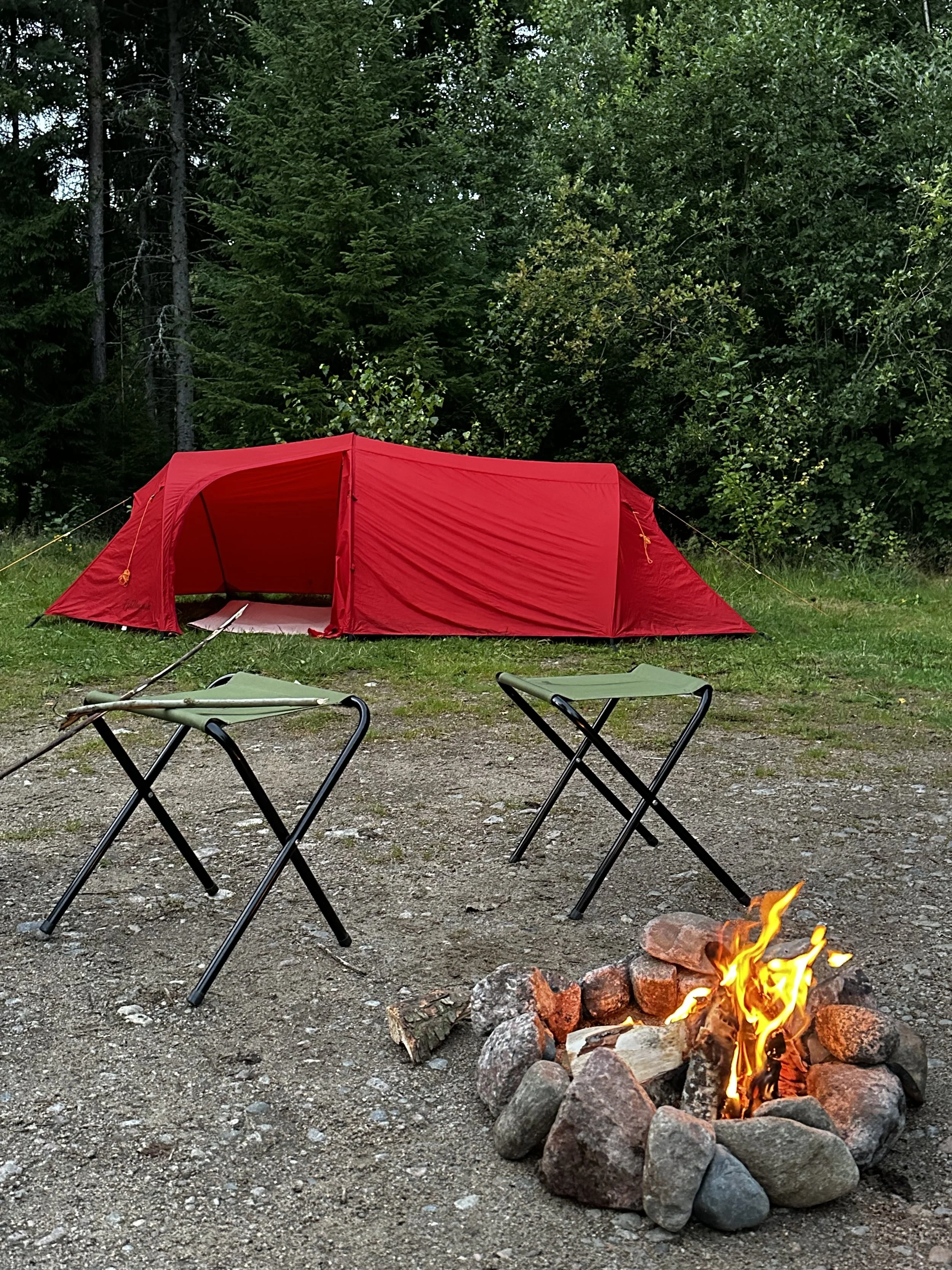 A fire burning at a fireplace with two green camping chairs next to it and a red tent in the background in a forest setting