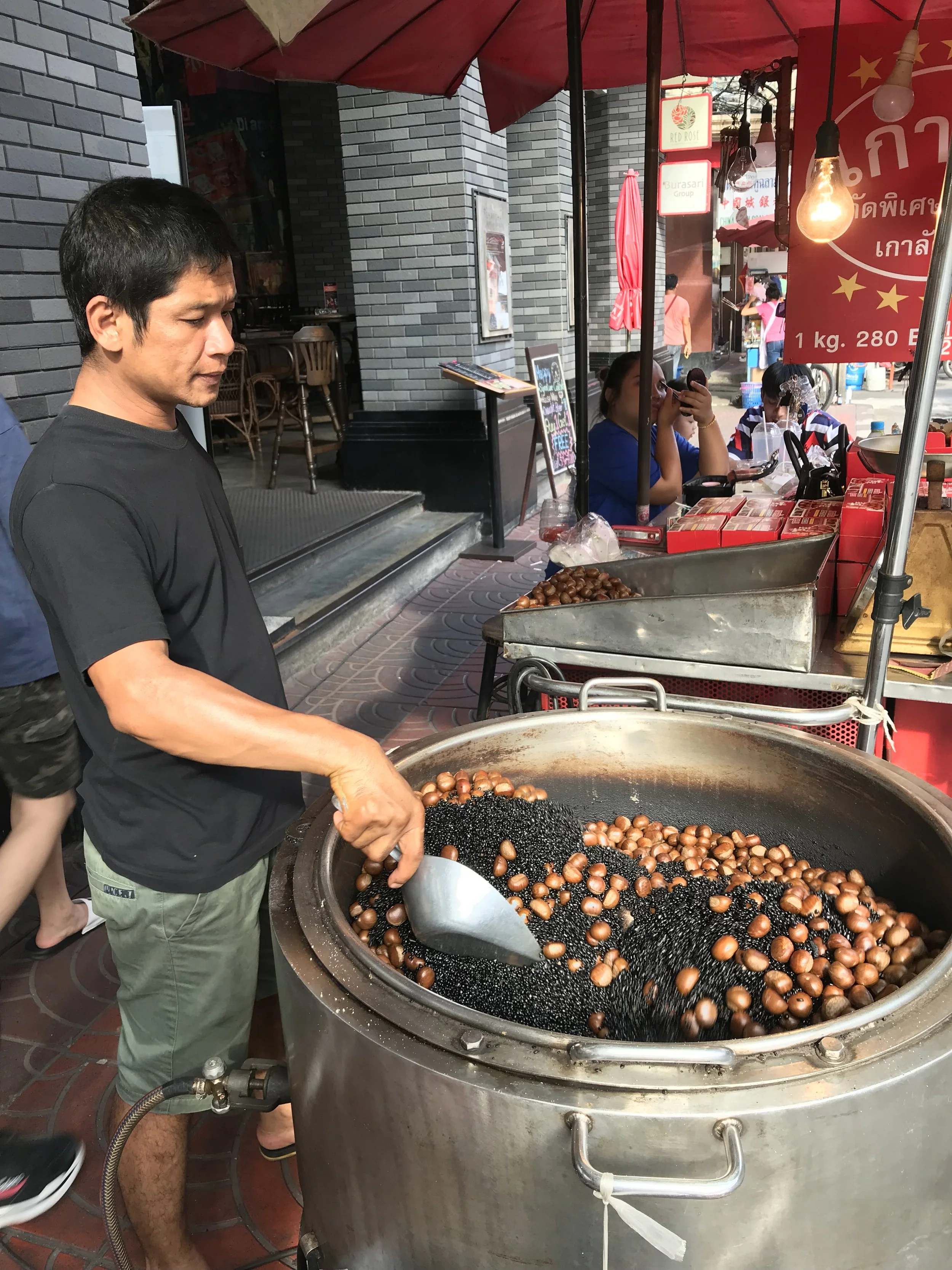 Man cooking chestnuts in a large metal wok on a street, using a metal scoop, with a market stall and people in the background.