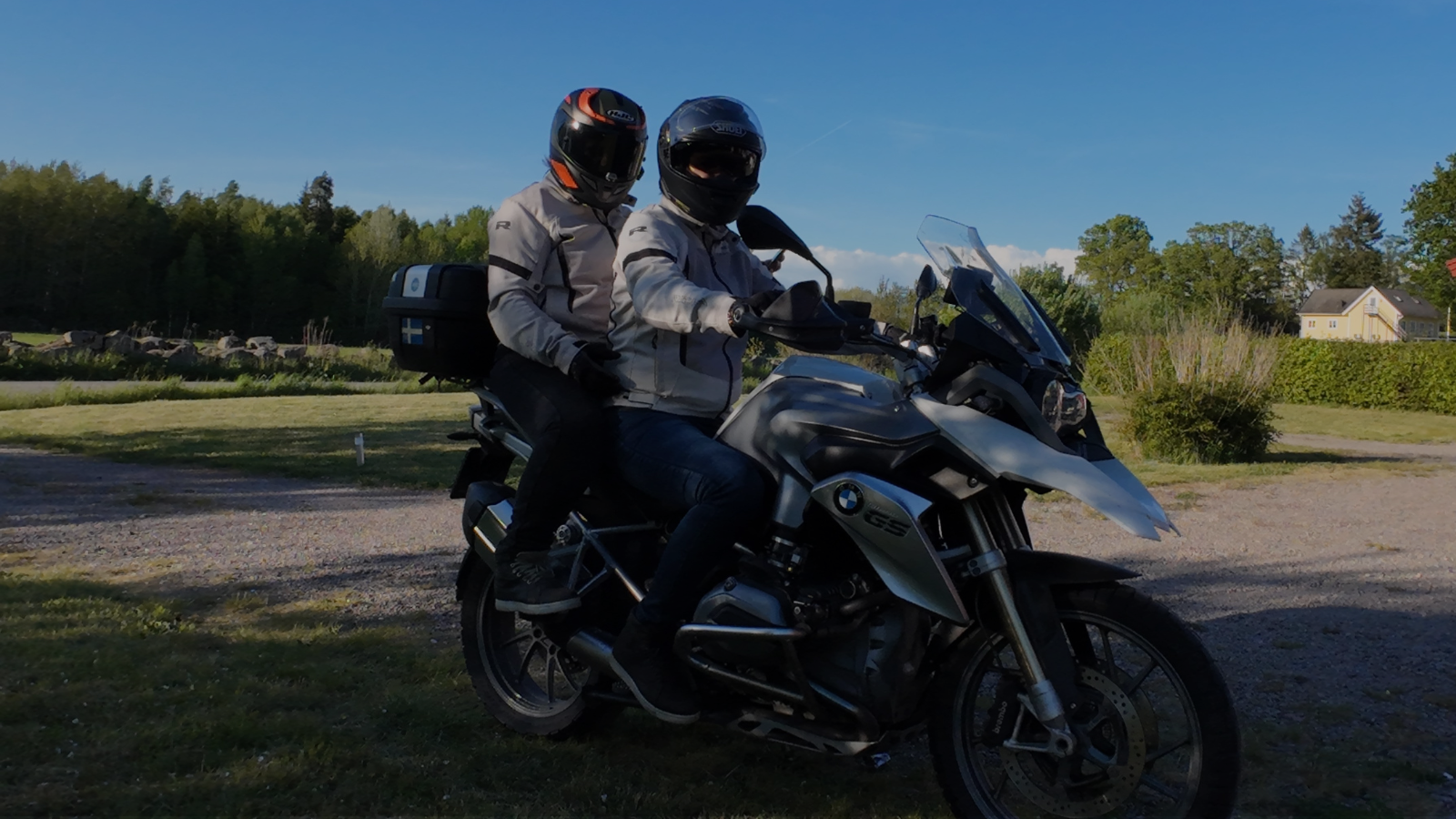 Two people wearing motorcycle helmets and jackets riding a BMW GS motorcycle on a rural road under a clear blue sky with trees and houses in the background.