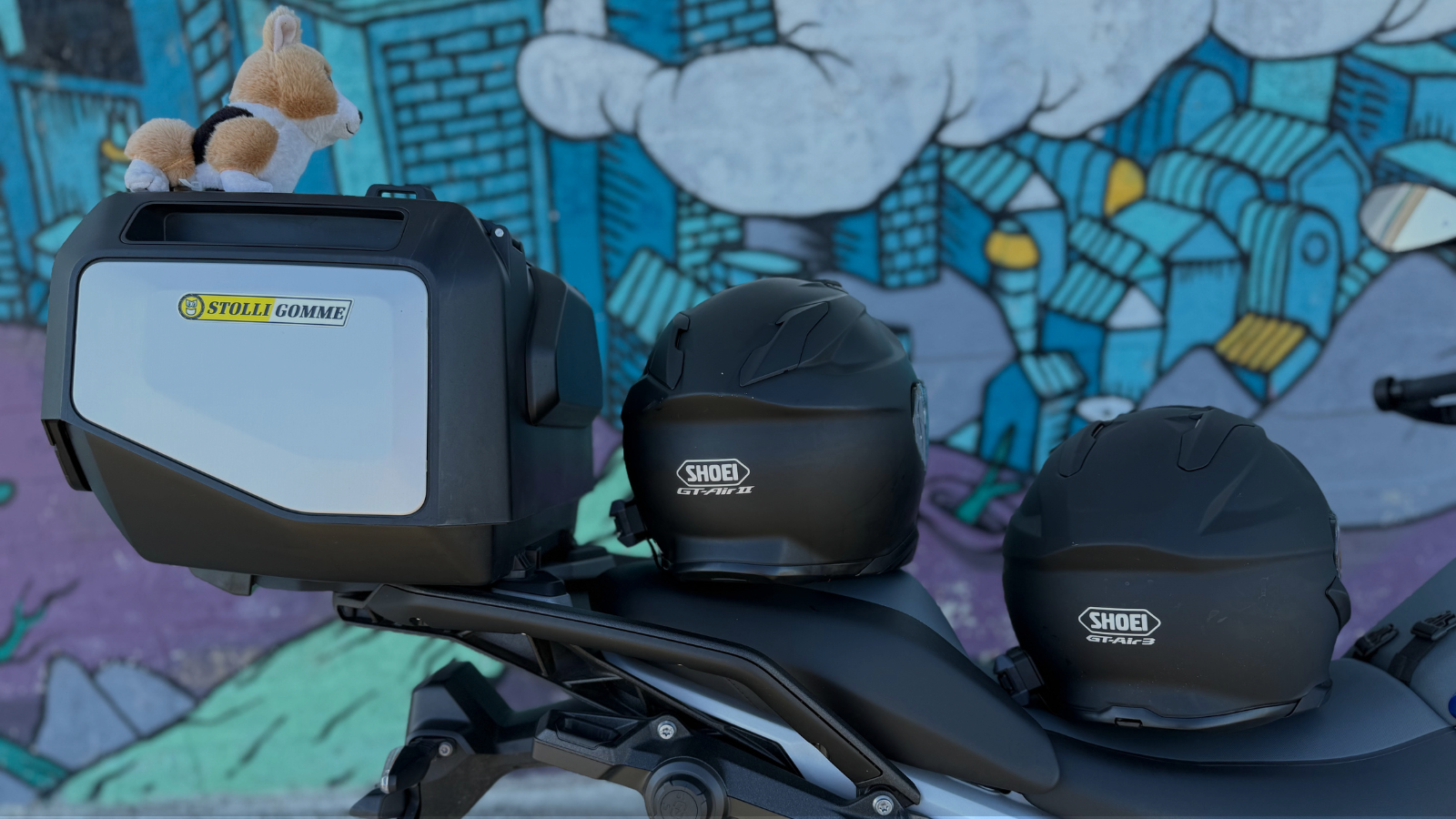 Motorcycle with two black Shoei helmets mounted on it, a stuffed dog toy on top of a grey and white Stolli Gomme storage box, against a colorful graffiti mural.