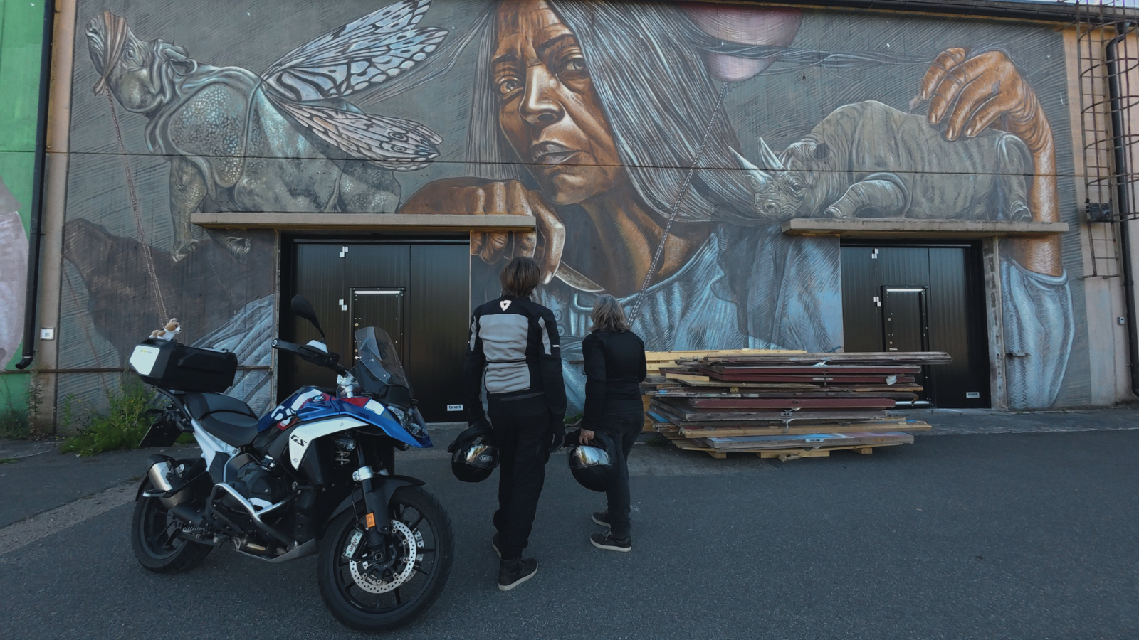 A mural on a building wall depicting a Native American woman with long hair, a dragonfly, a rhinoceros, and a hand. Two people with motorcycle helmets watch the mural, and a motorcycle is parked nearby.