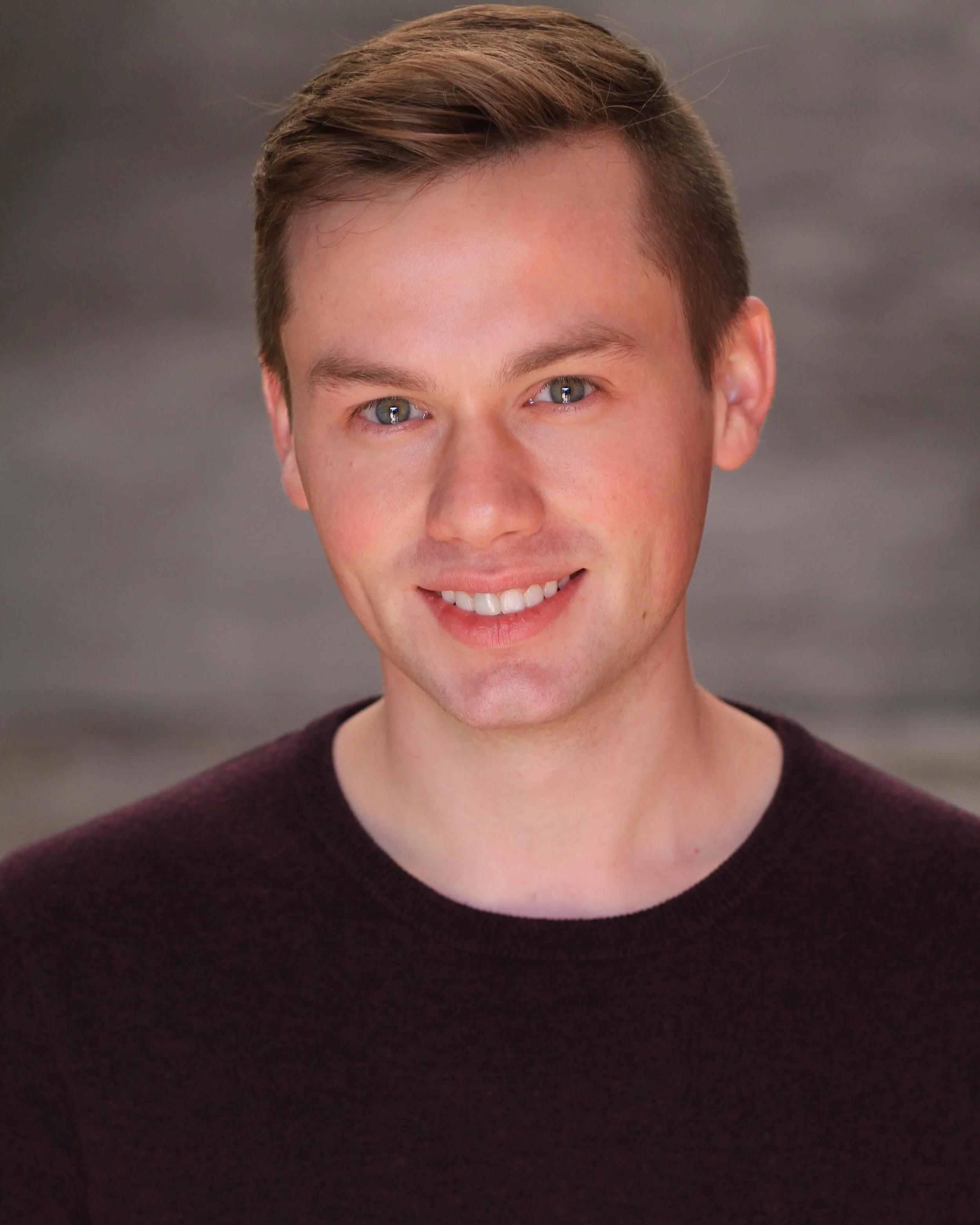 A young man with short brown hair, blue eyes, and a friendly smile, wearing a dark maroon sweater, standing against a neutral background.