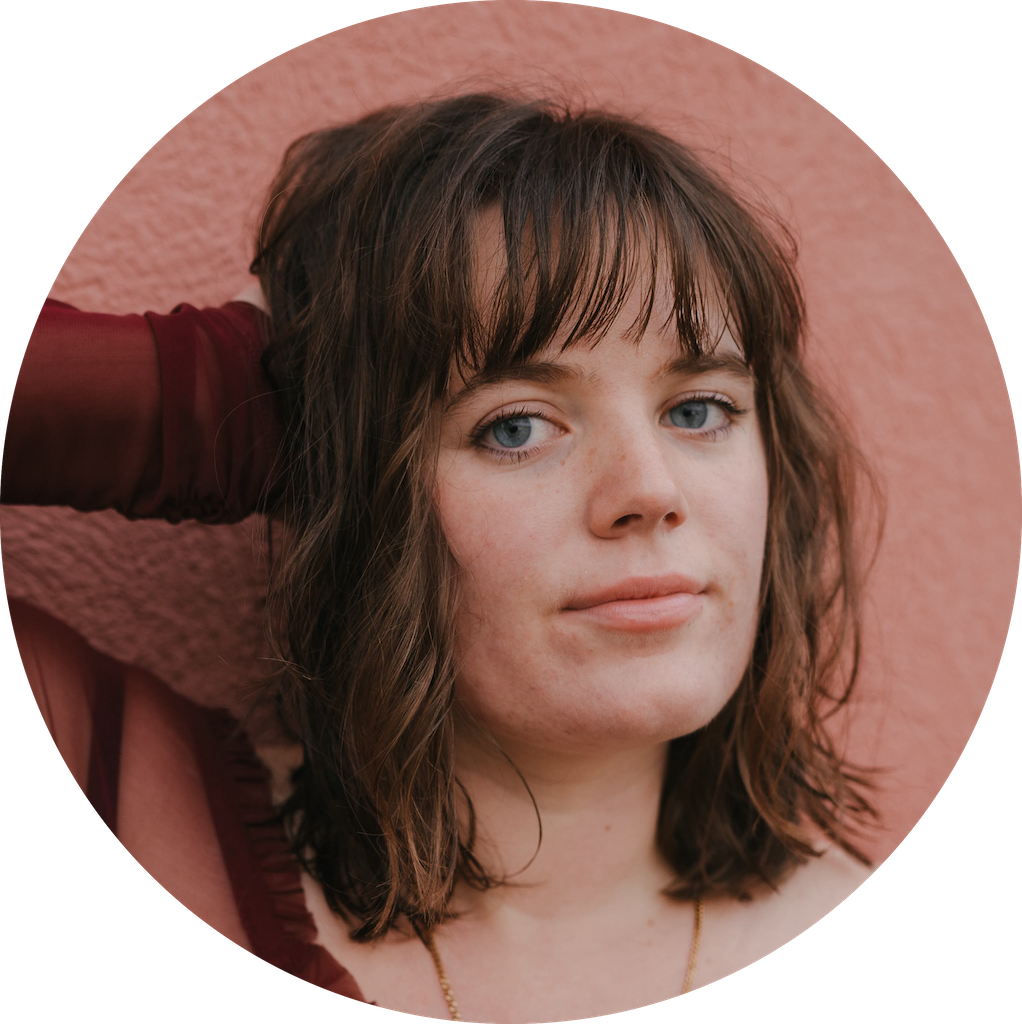 Close-up portrait of a woman with blue eyes, shoulder-length brown hair with bangs, and light skin, standing against a pink textured wall.