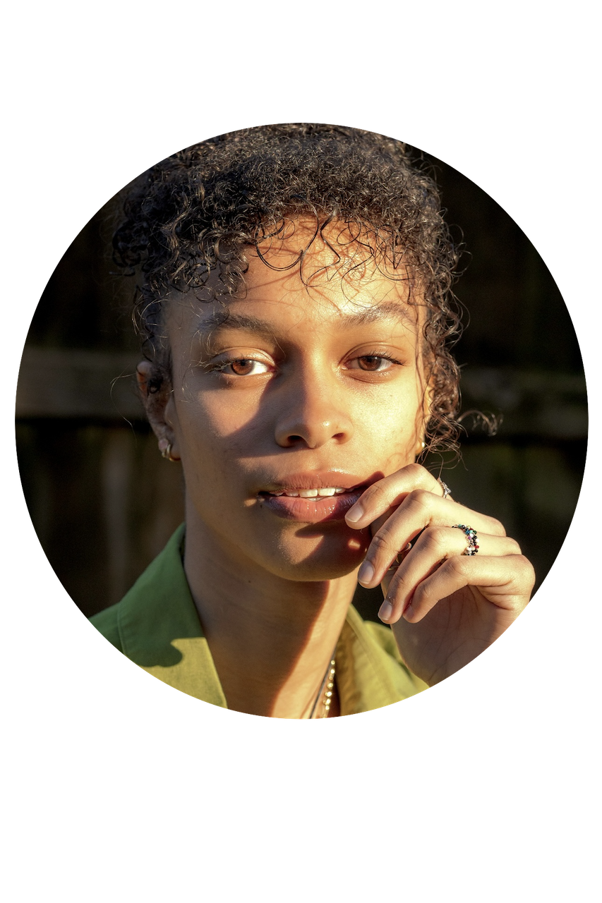 Close-up of a young woman with curly hair, wearing a green shirt, touching her chin with her hand, outdoors during golden hour.
