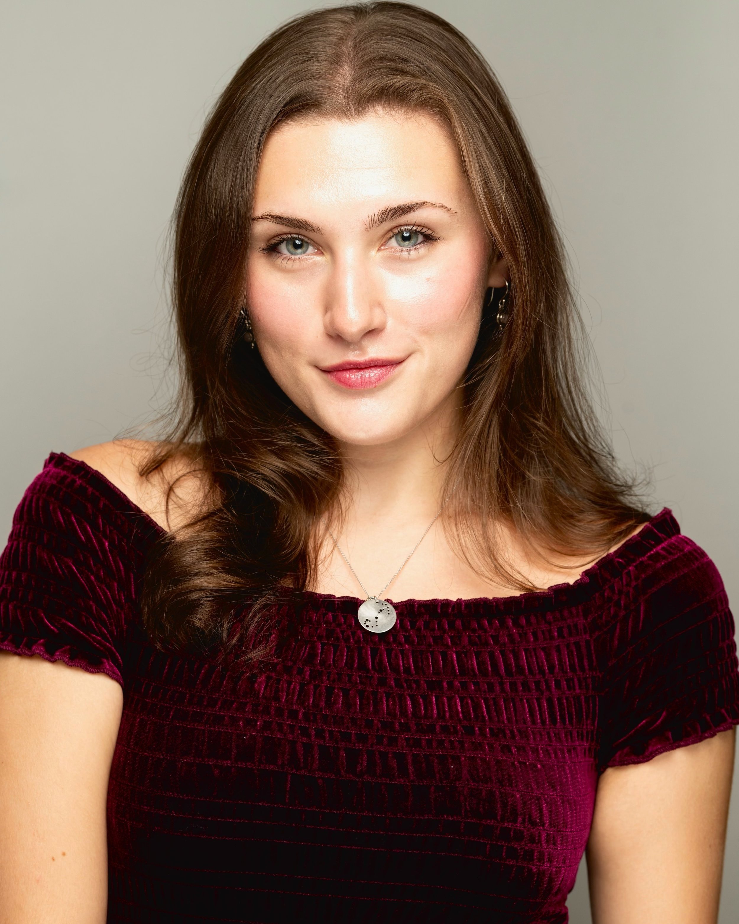 A young woman with shoulder-length brown hair, blue eyes, and light skin, wearing a burgundy velvet top and a silver necklace with a round pendant. She has a slight smile and is looking directly at the camera.