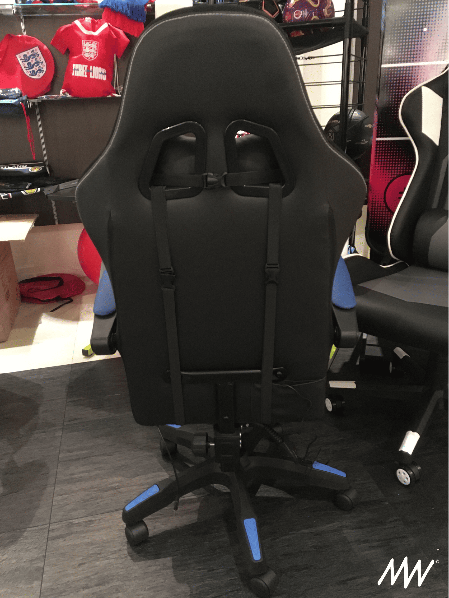 A black gaming or office chair with blue accents, positioned in front of shelves with sports merchandise and other gaming chairs.
