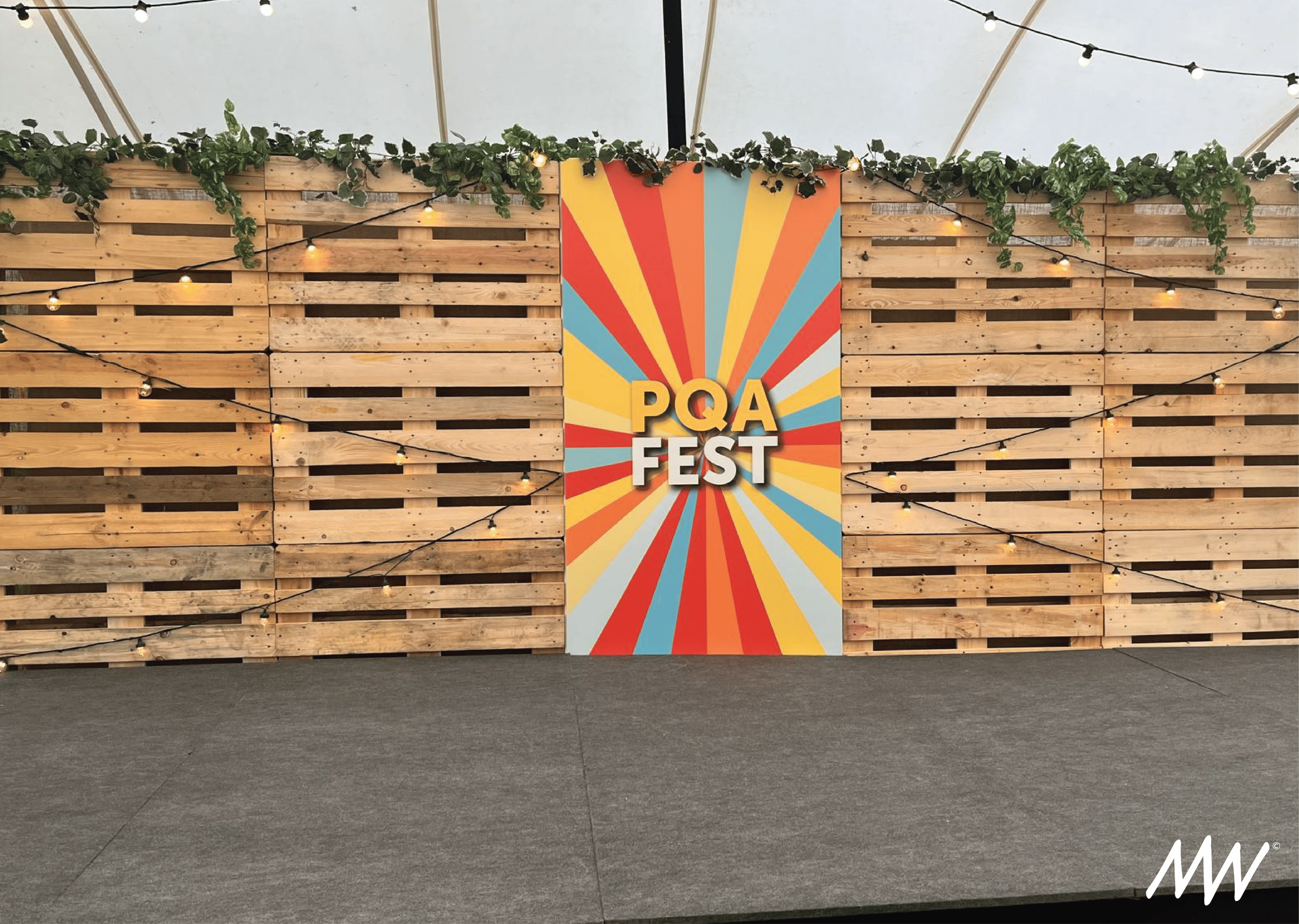 Wood pallet backdrop decorated with string lights and green foliage, featuring a colorful vertical banner that reads 'PQA FEST' with a sunburst pattern in red, yellow, blue, and gray.