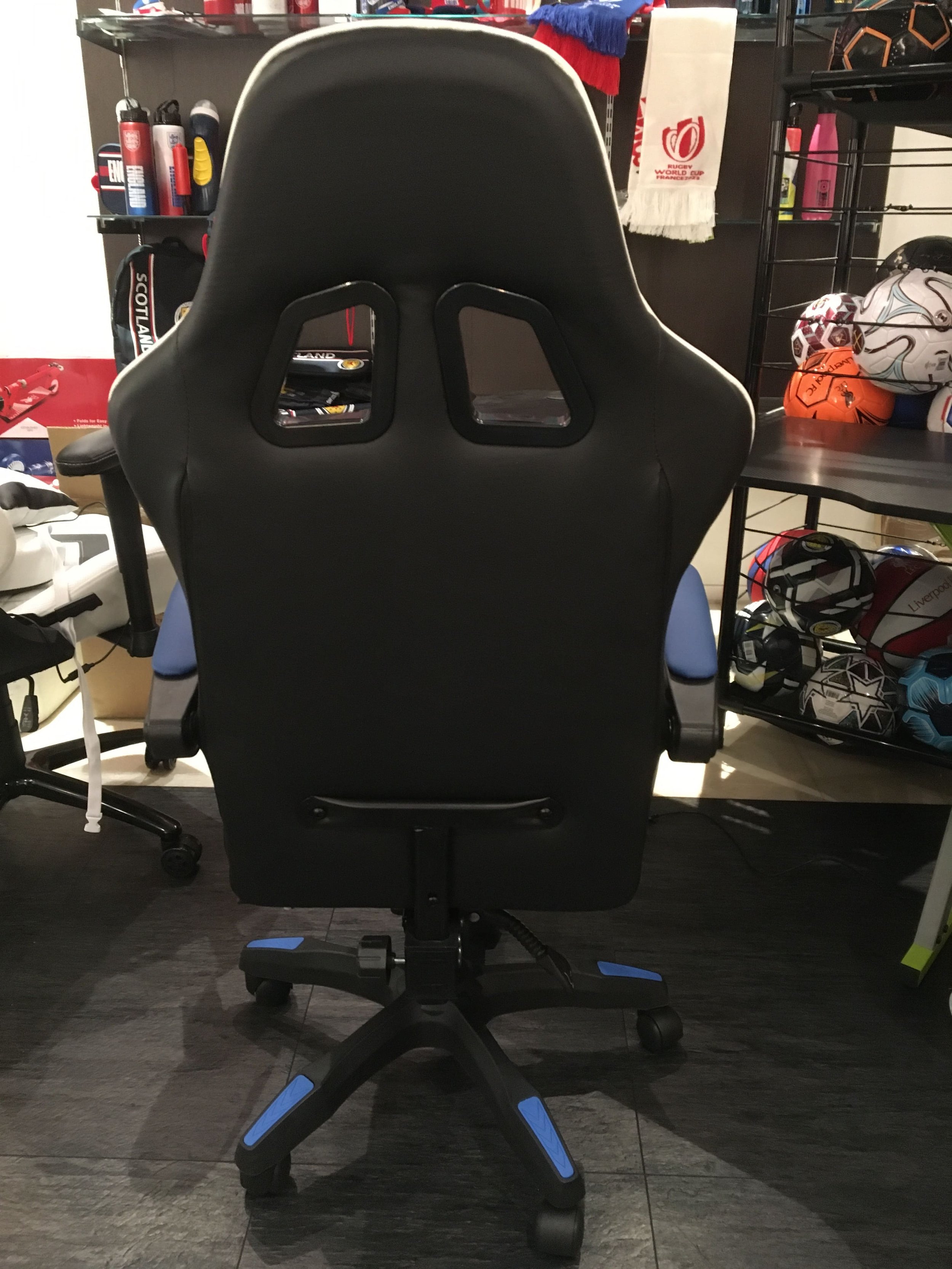 A black gaming or office chair with a high back and two cut-out holes near the top, situated in a room with sports equipment and shelves around.