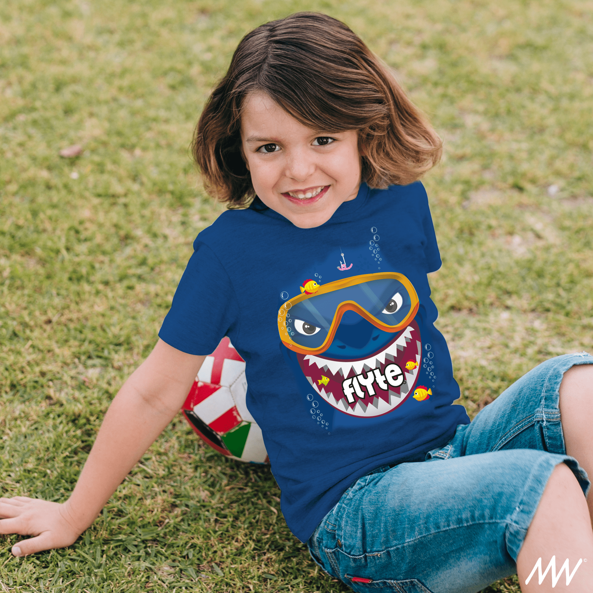 Young boy with brown hair smiling, sitting on grass, wearing a blue T-shirt with a shark face design, the word 'FYTE' on it, and small colorful fish, with a soccer ball nearby.