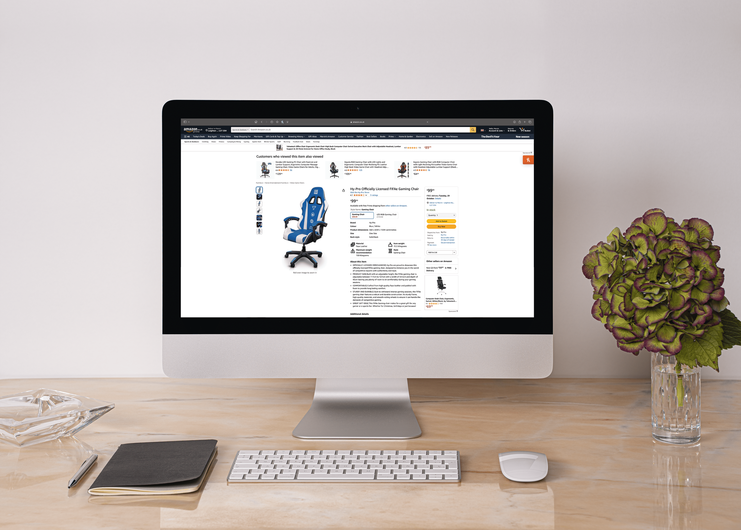 A computer monitor on a wooden desk displaying an online shopping website with a blue gaming chair. There is a black notebook, a pen, a white keyboard, a white mouse, and a glass vase with green and purple leaves on the desk. The background is plain white.