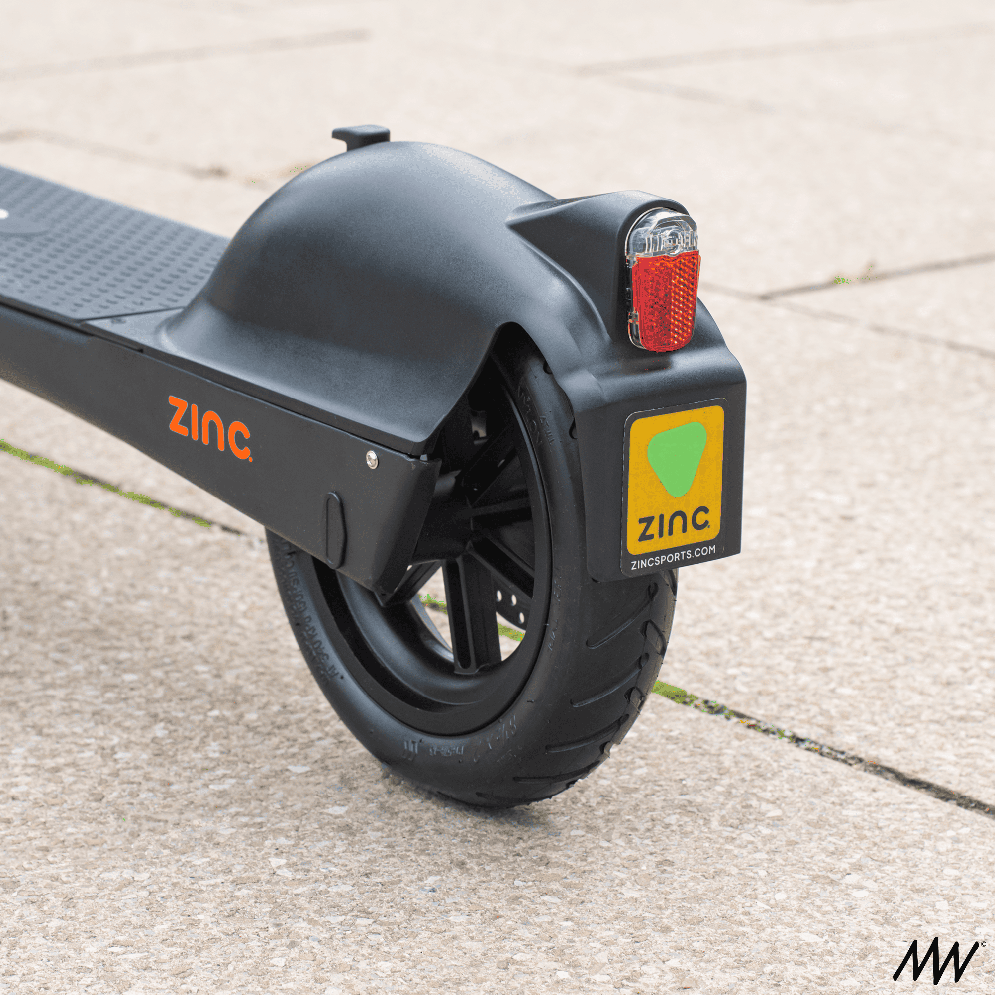 Close-up of a black ZINC electric scooter rear wheel with a yellow ZINC logo on a mudguard, showing a red taillight and a license plate with green and yellow logo.