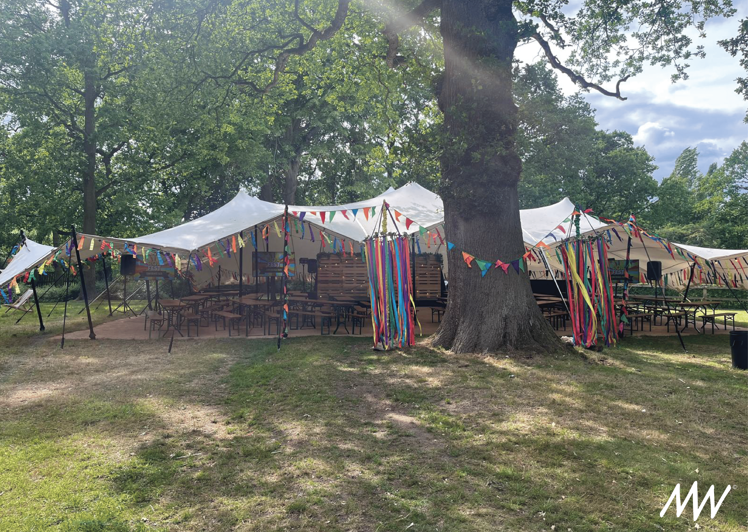 Outdoor event setup with large white tents decorated with colorful pennant banners and streamers, situated among large trees on a grassy area.