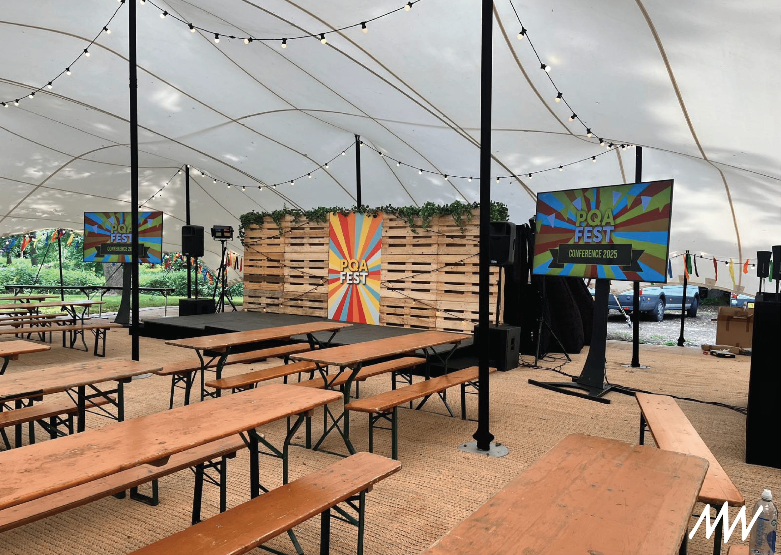 Indoor event space with wooden picnic tables, string lights, a stage with a wooden pallet backdrop decorated with greenery, and two large screens displaying "PQA FEST Conference 2025" at a festival.