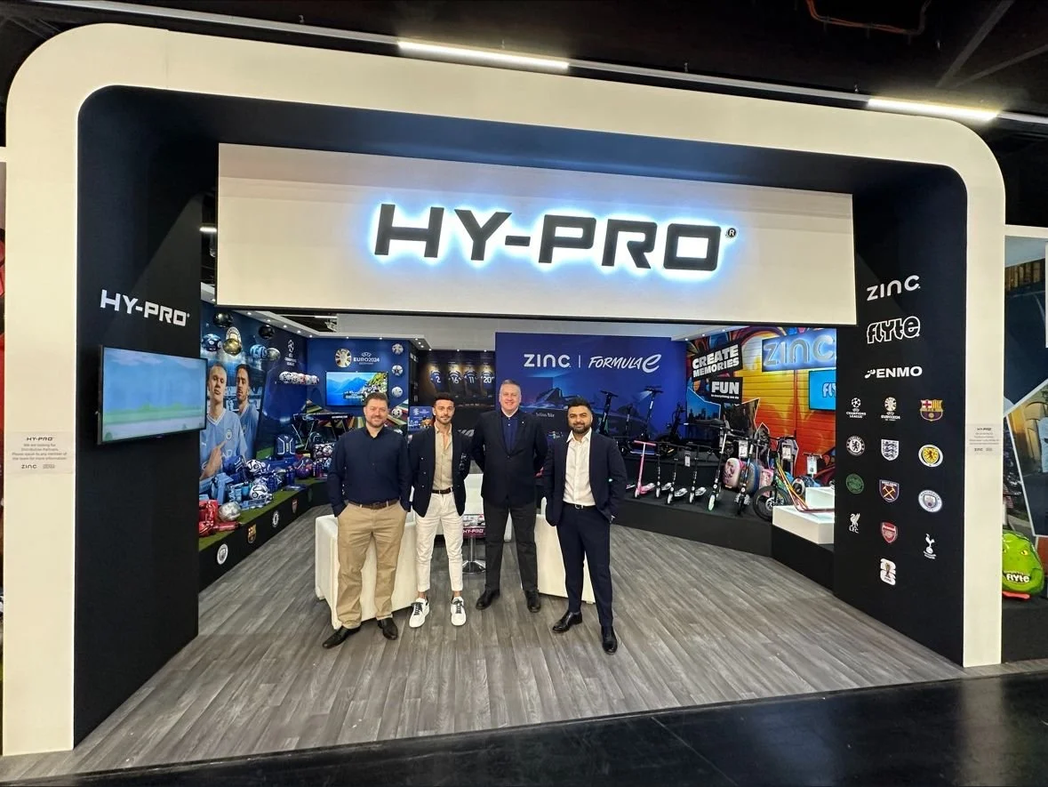 Four men standing in front of a Hy-Pro booth at an event with sports merchandise, bikes, scooters, and a sports-themed display in the background.