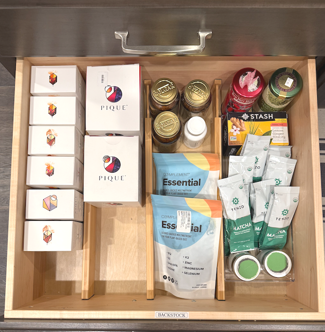 Open kitchen drawer containing dietary supplements, herbal teas, and vitamins. Items include white boxes with geometric logos, bottled drinks, supplement jars, and herbal tea packets labeled 'Matcha' and 'Lemon Ginger'.