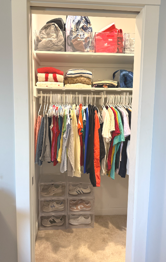Open closet with hanging clothes, storage bins with shoes underneath, and shelves with bags and folded towels.