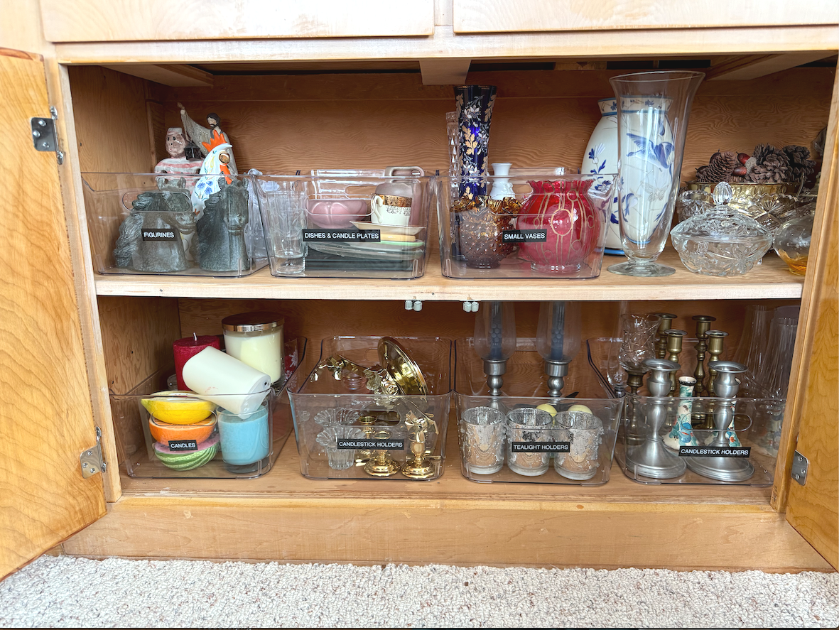 A wooden cabinet with three shelves containing various decorative and household items, including figurines, dishes, candle plates, small vases, candles, candlestick holders, and tealight holders.