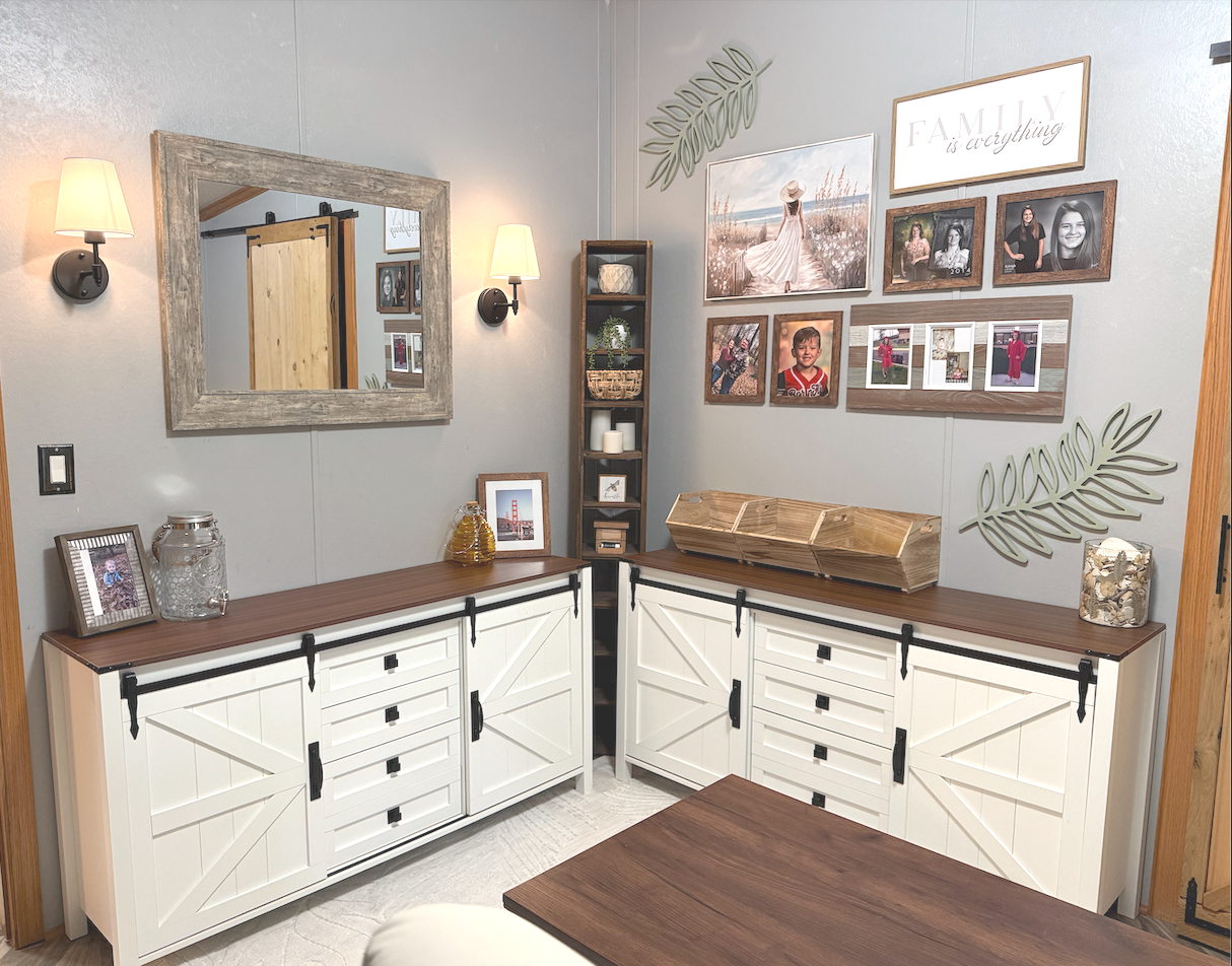 A corner of a room with white cabinets with black hardware, a wooden countertop, and decorative items including picture frames, jars, and a wooden tray. The wall has framed photos, family quotes, and leaf-shaped wall decor, with two wall sconces and 