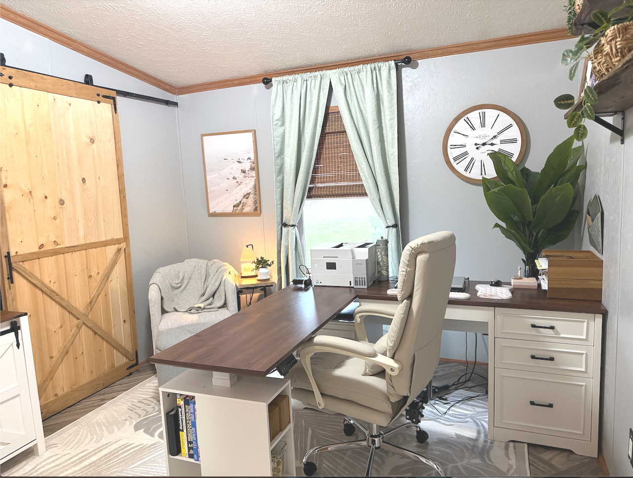 Home office with a wooden sliding barn door, a large clock with Roman numerals, a window with light-colored curtains, a white desk with an office chair, a printer, and a large potted plant.