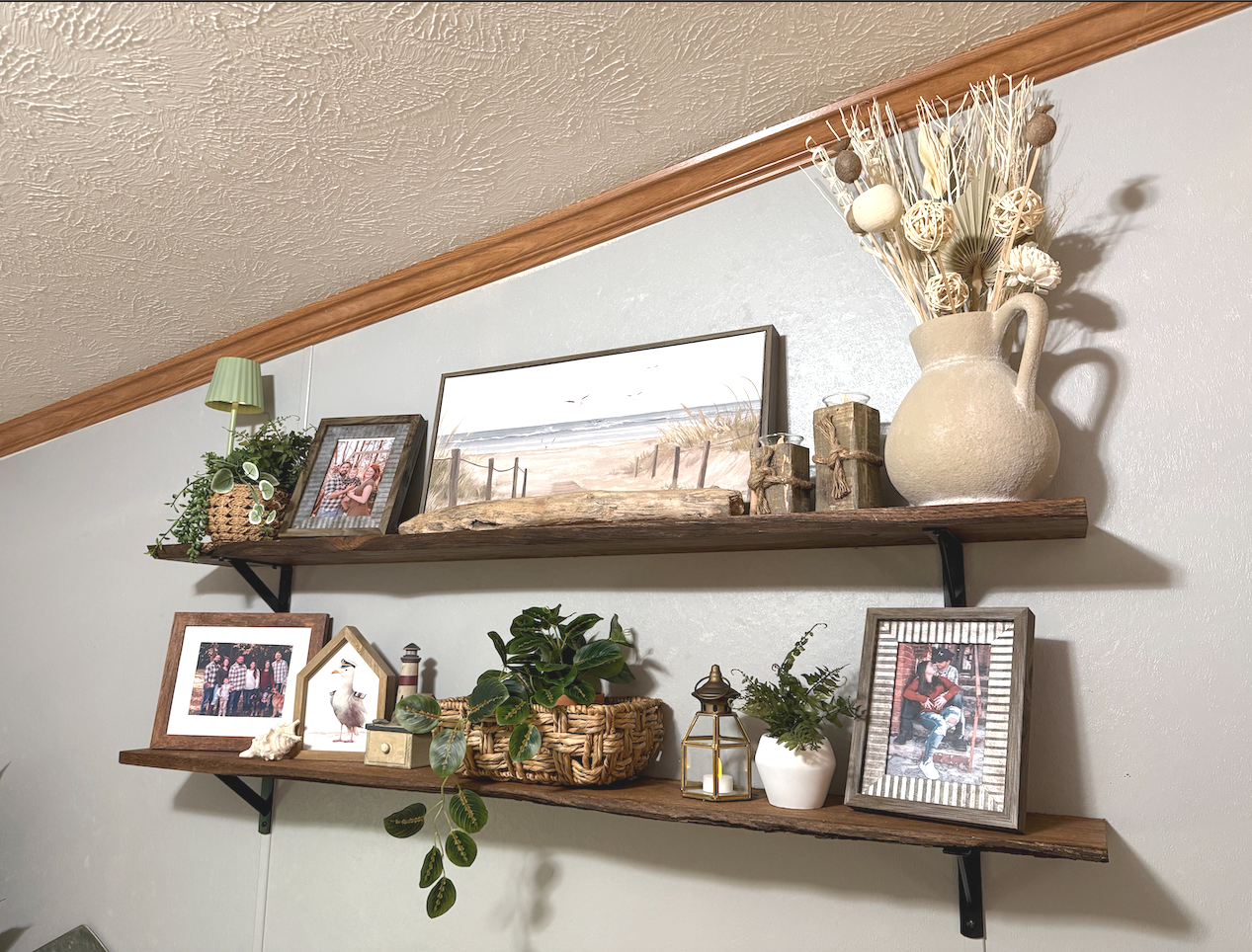 Two wooden shelves with decorative items including framed photographs, potted plants, a lantern, candles, and a large ceramic vase with dried flowers, mounted on a light-colored wall.