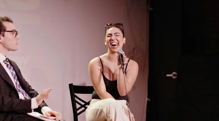 A woman laughing and holding a microphone during a panel or interview, with a man sitting beside her looking at her.
