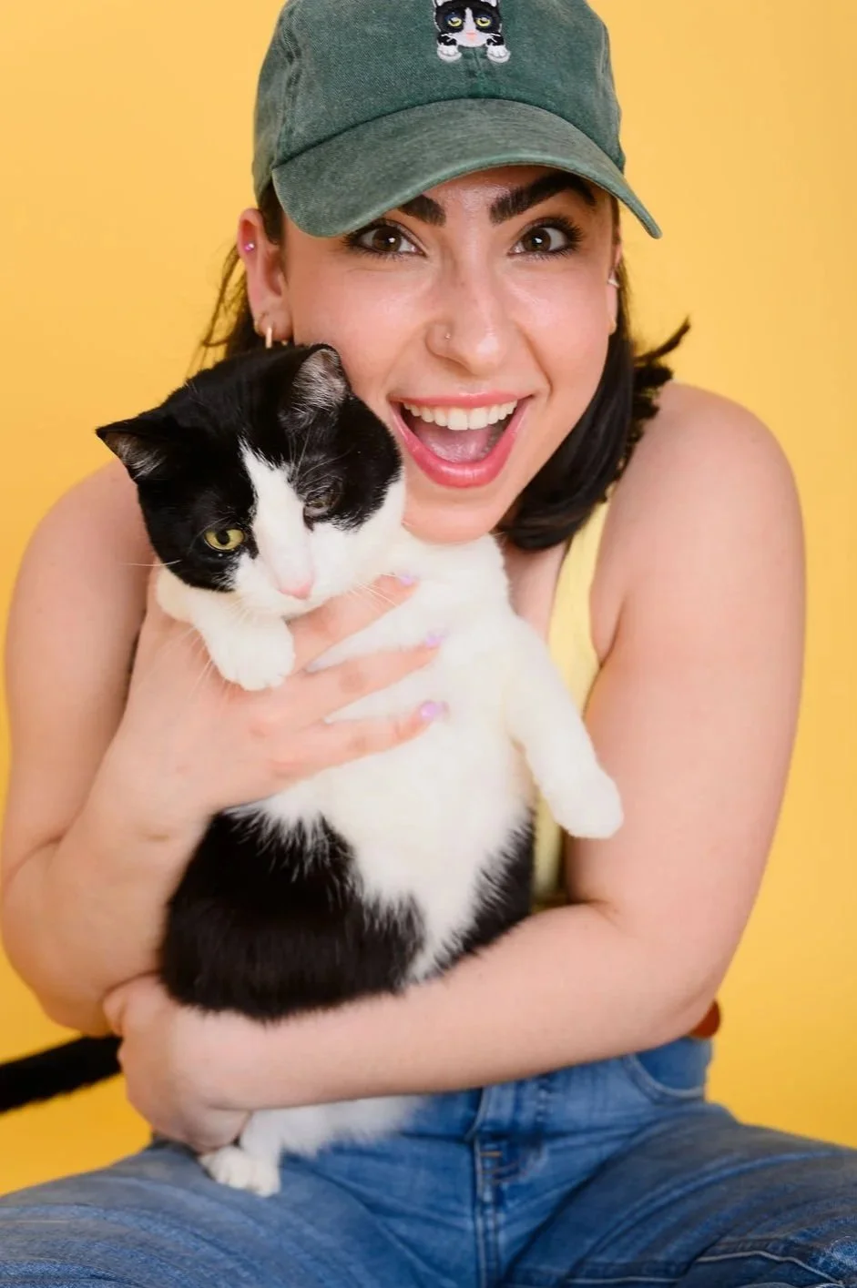A young woman with a green baseball cap smiling and holding a black and white cat against a yellow background.