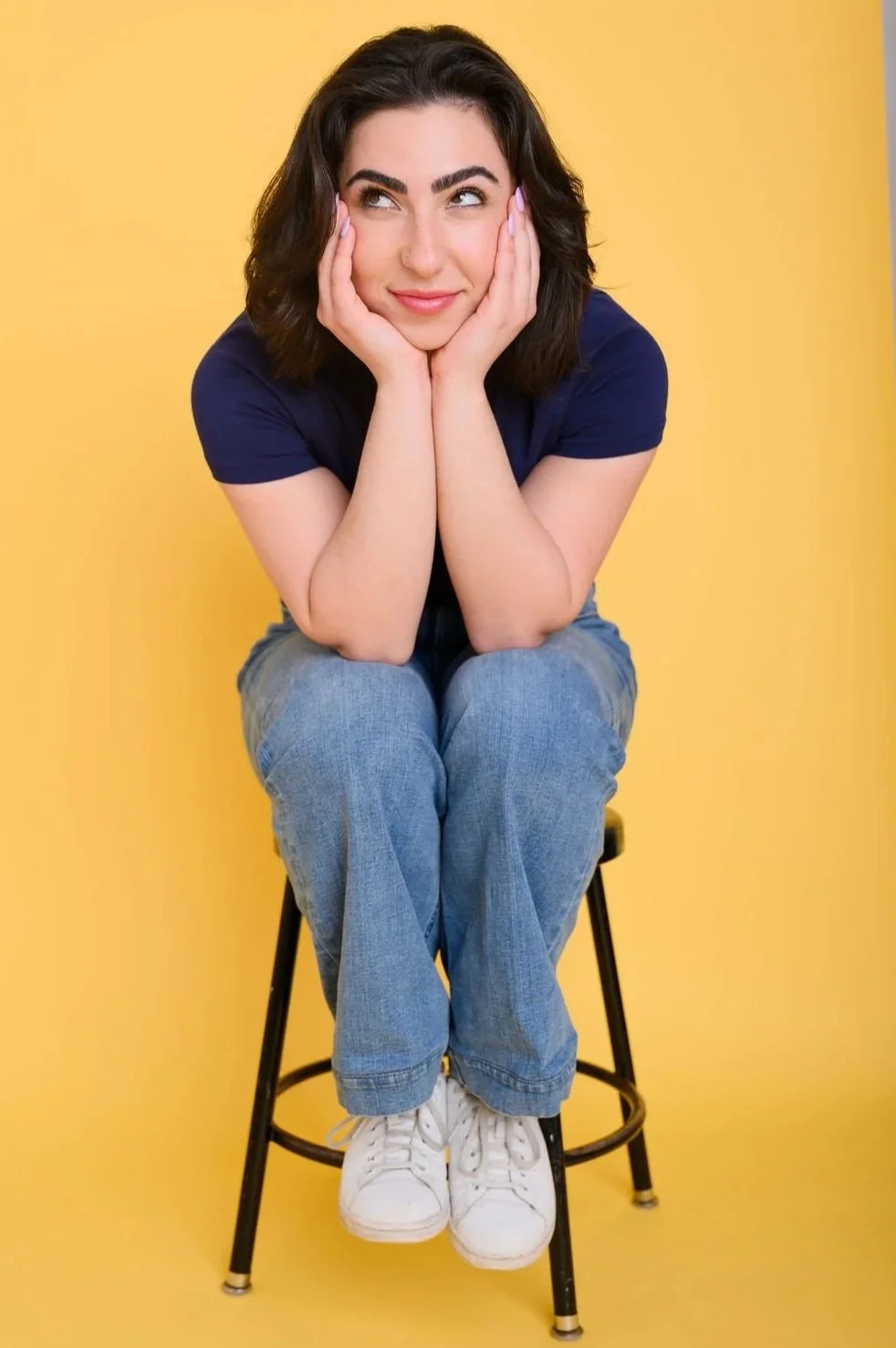 A woman with dark wavy hair smiling, resting her chin on her hands, sitting on a stool against a yellow background, wearing a navy t-shirt, jeans, and white sneakers.